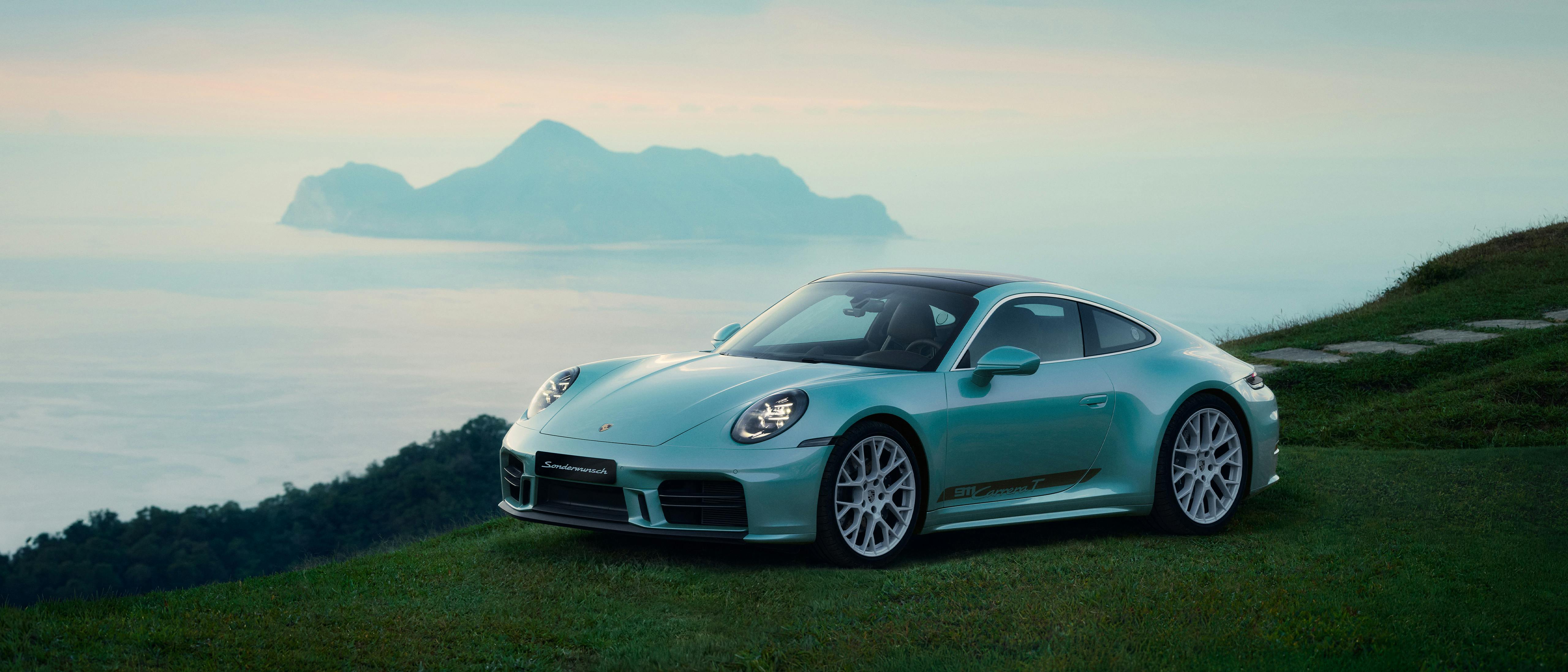 A light blue Porsche 911 Carrera T parked on a grassy hill overlooking the sea and distant islands at sunset.