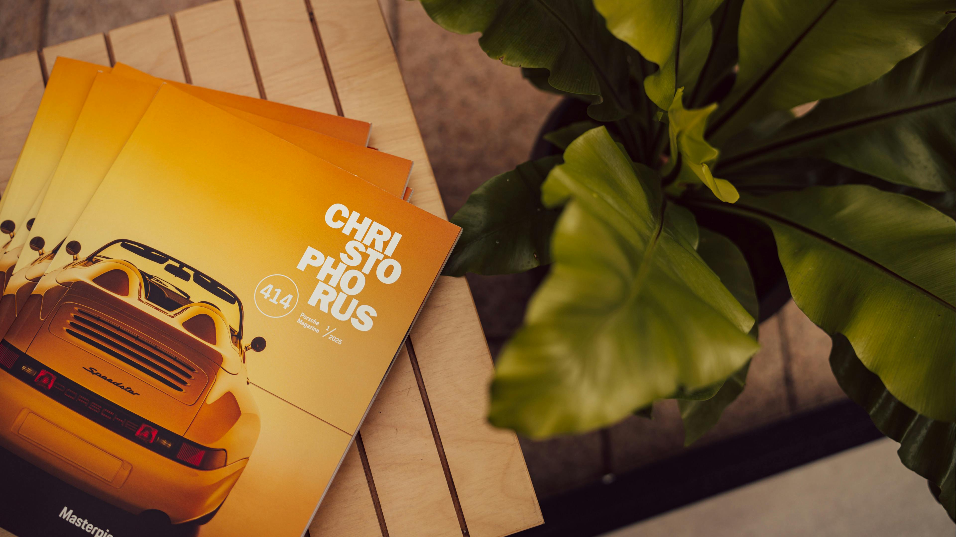 A top-down view of a stack of Christophorus Porsche magazines resting on a light wood slatted table. The magazine cover features a classic yellow Porsche Speedster.