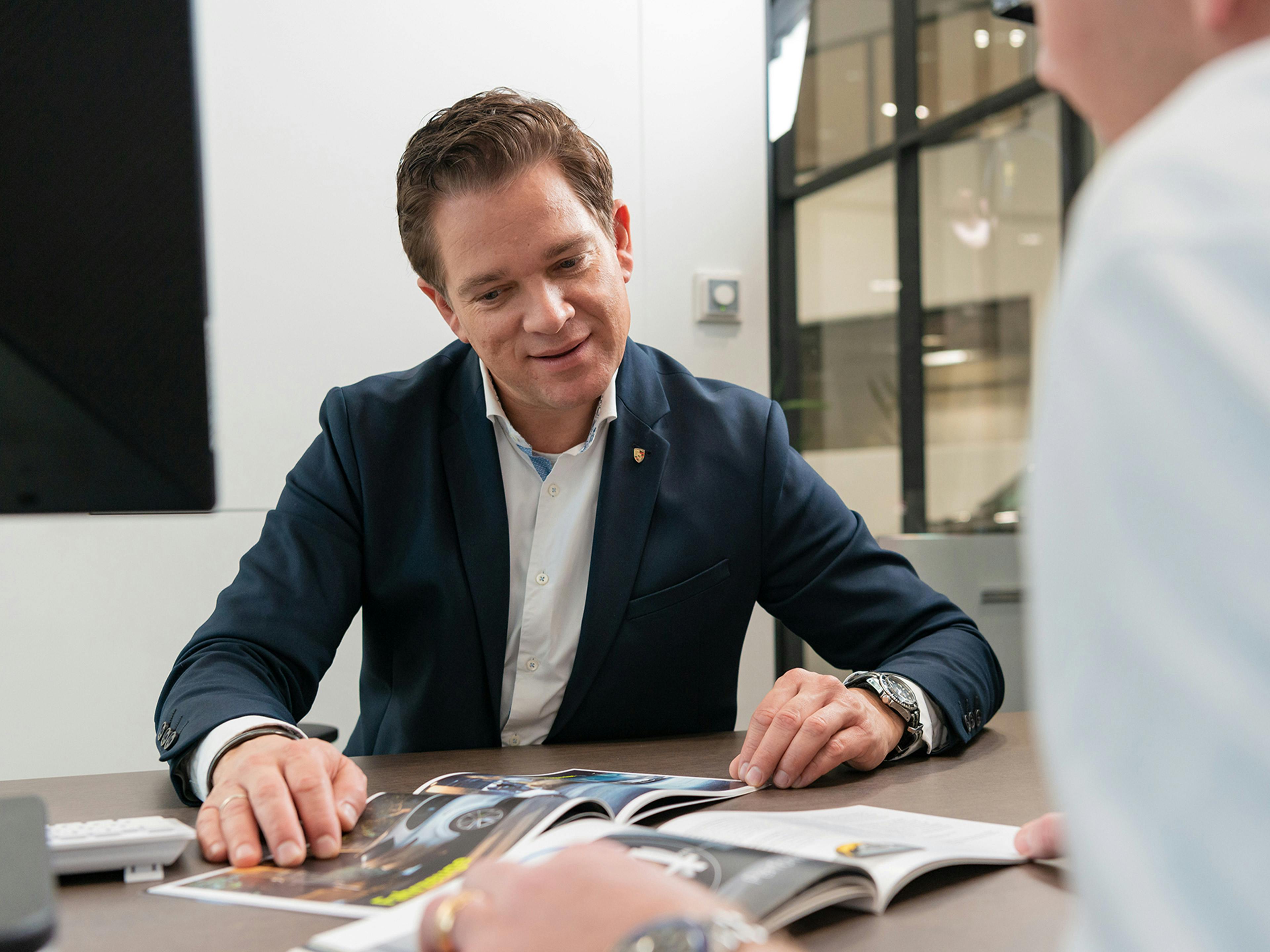 Un collaborateur de Porsche assis derrière un bureau dans une salle de réunion montre à un collègue masculin quelque chose dans un catalogue posé sur le bureau entre eux.