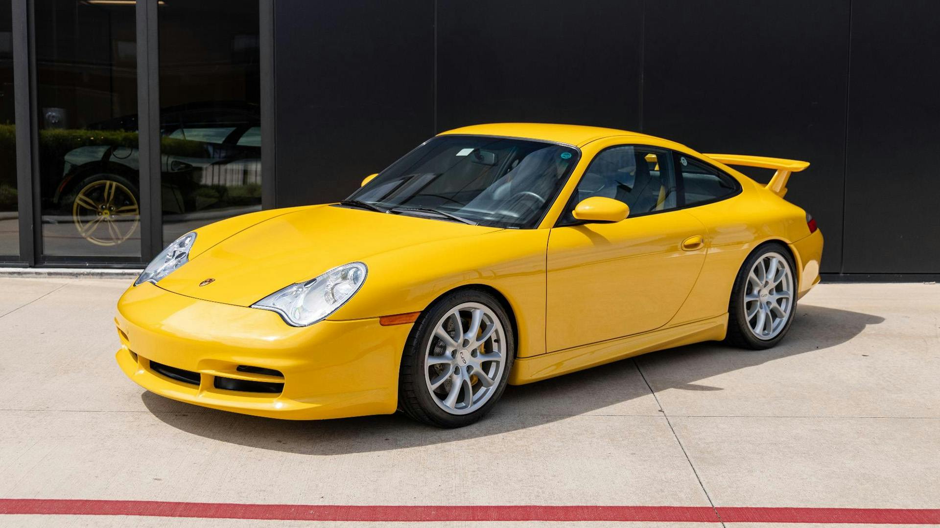 A sleek, yellow 911 sports car with a spoiler is parked on a concrete surface near a black wall.