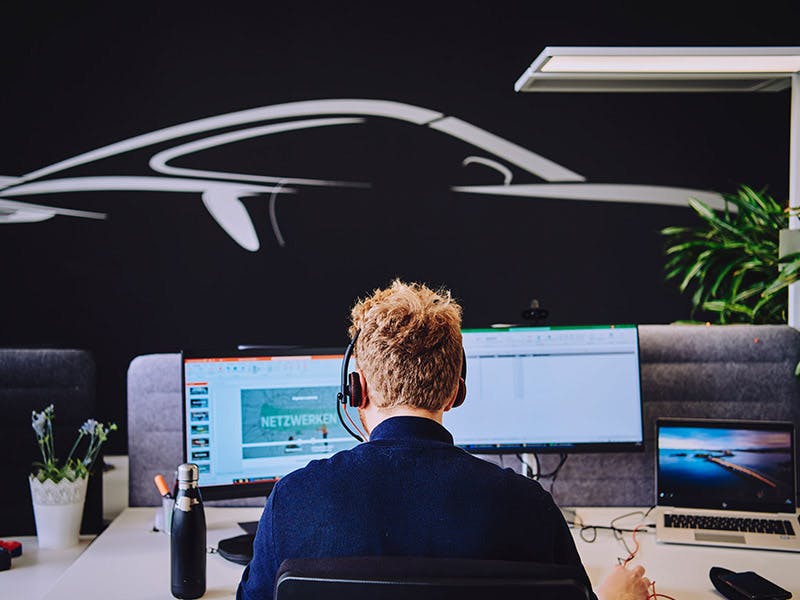 A student is working on his thesis at Porsche. He is sitting in front of two screens and a laptop.