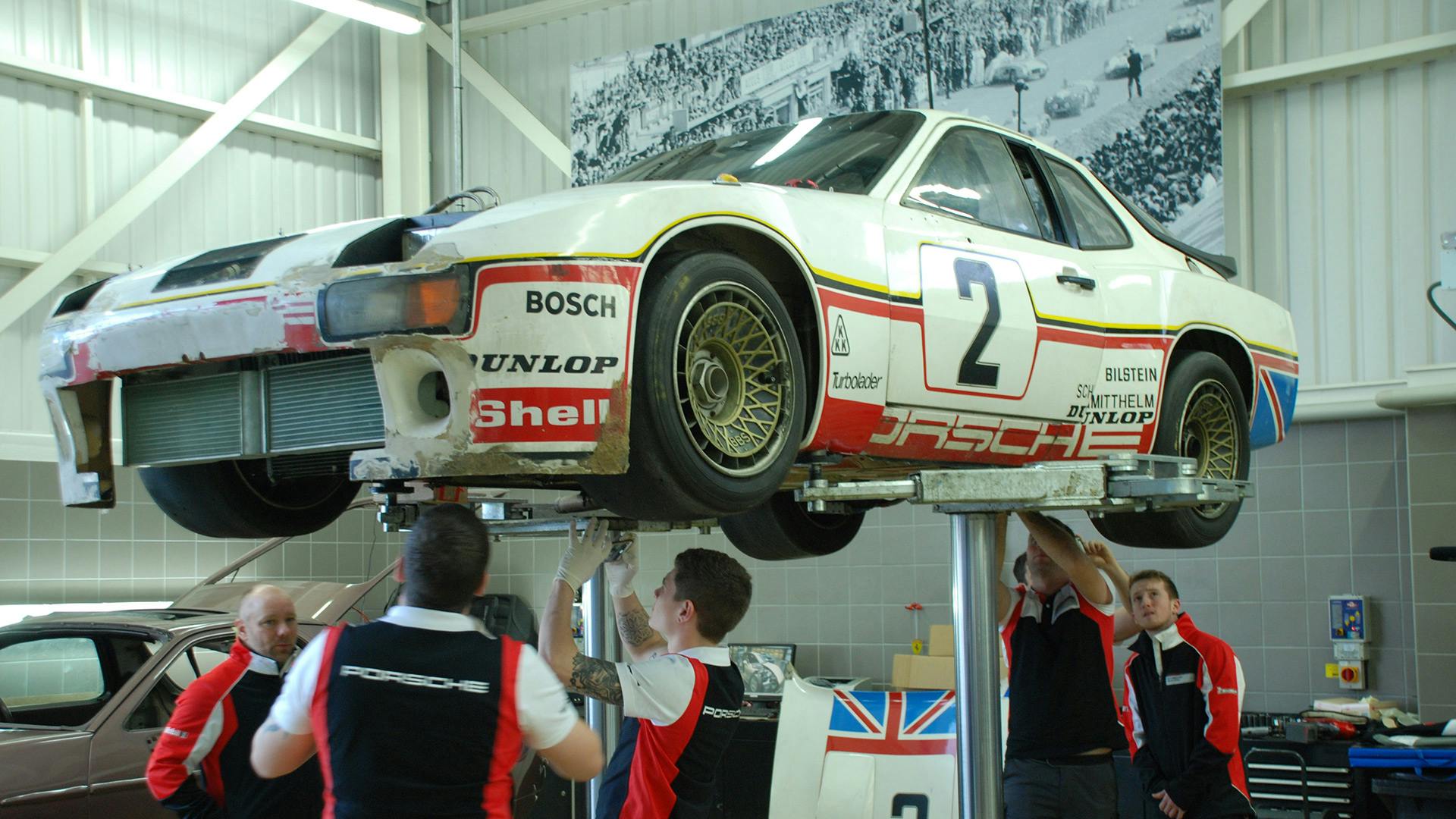 Multiple employees working on the bottom side of the Porsche 924 GTP which is standing on a lifting platform.