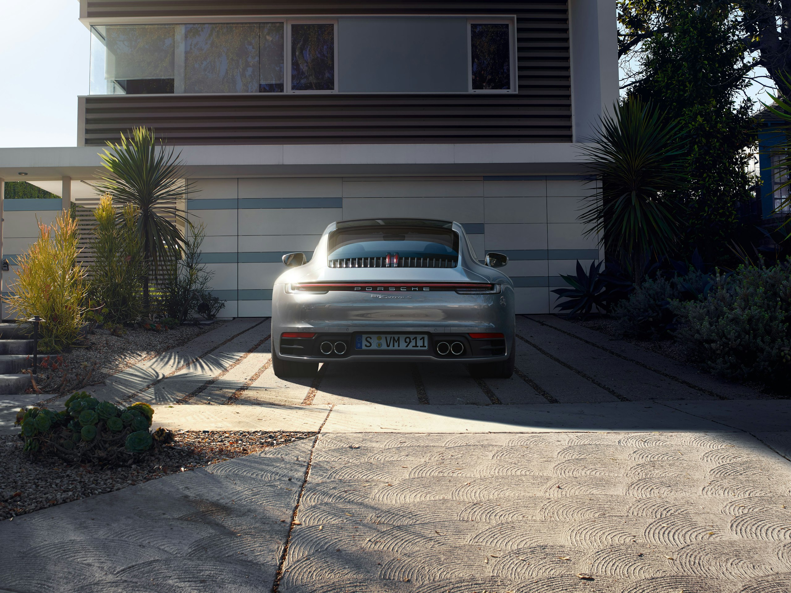 Rear shot of an anthracite 911 Carrera S parked in front of a modern residential building next to palm trees in the sunset
