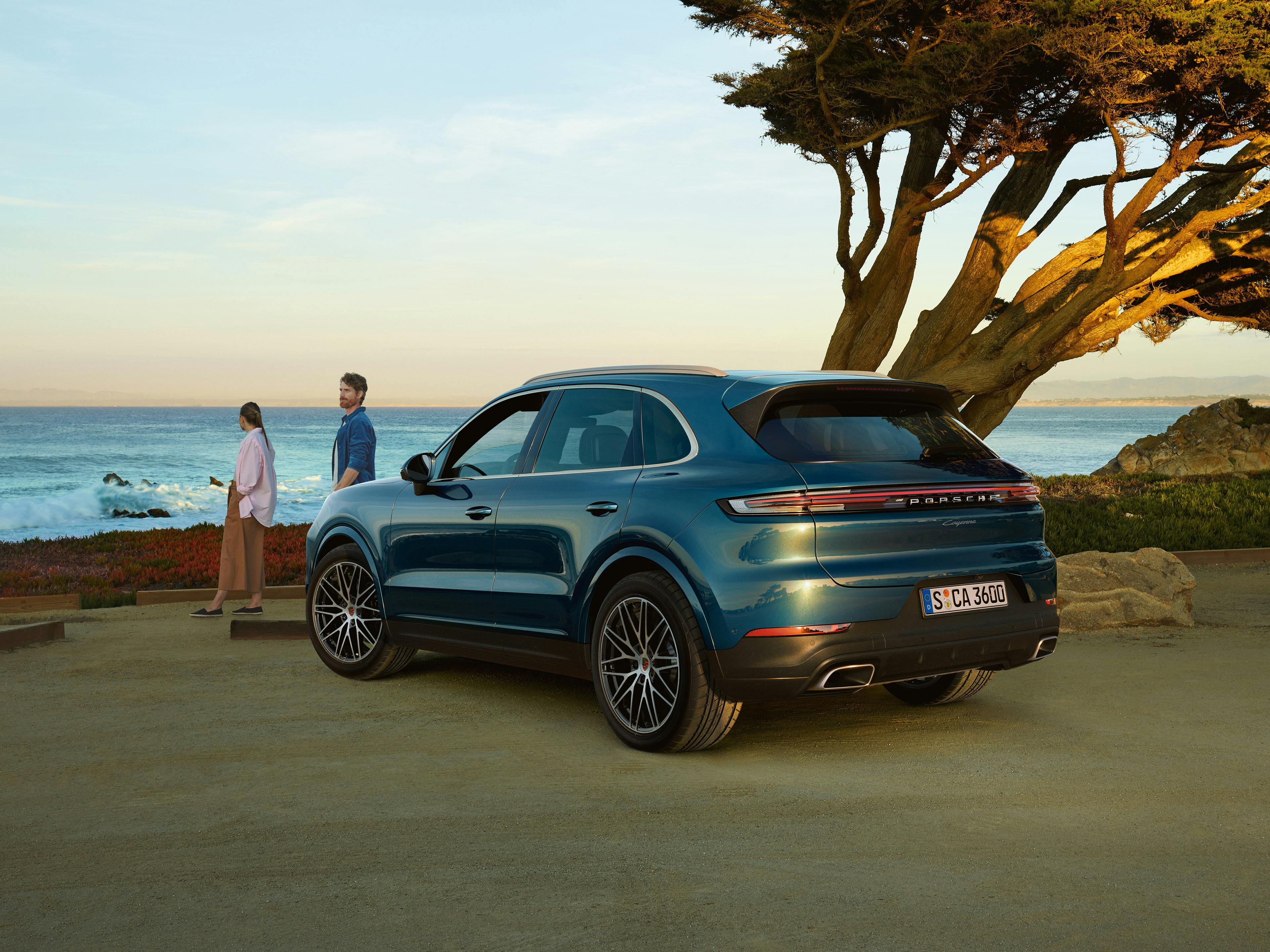 A blue Porsche Cayenne parked near a coastal road with two people standing by the ocean at sunset.