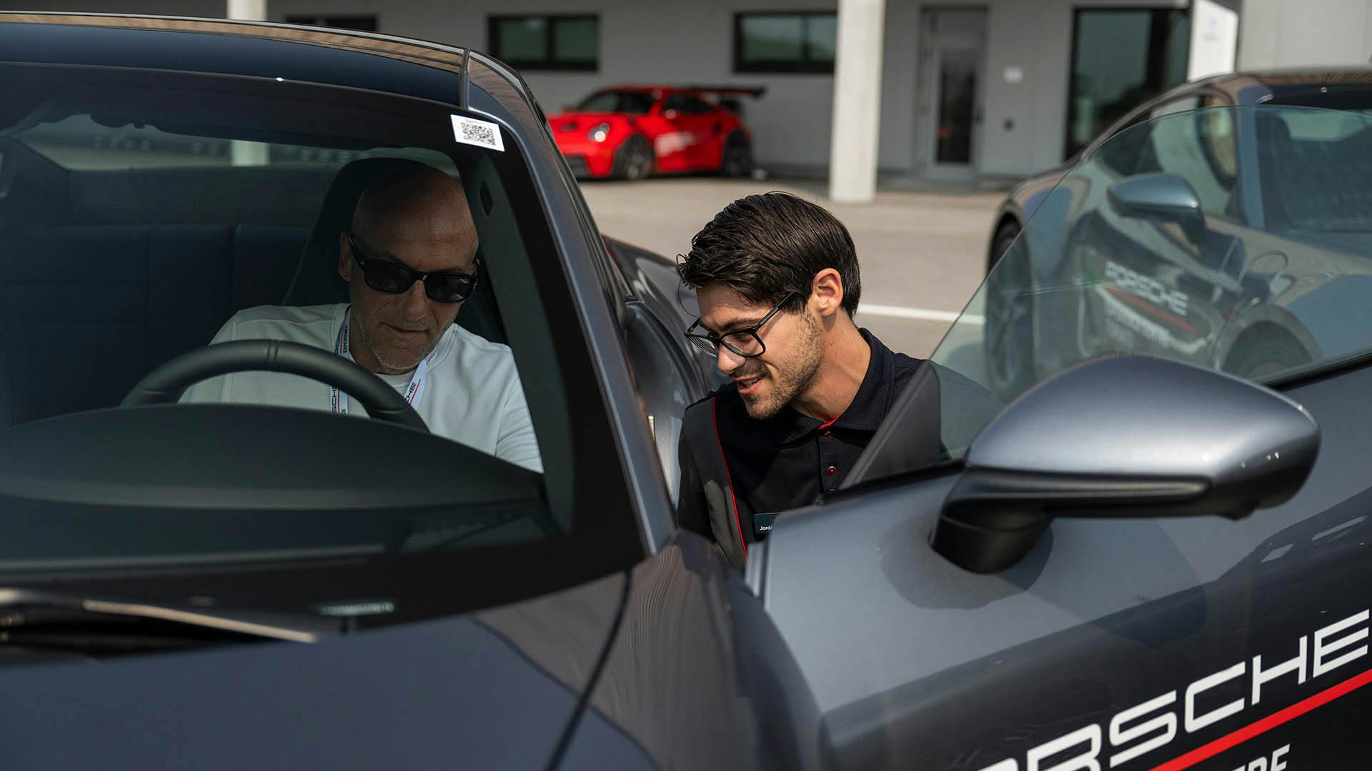Un moniteur accroupi à côté d'un conducteur assis dans une Porsche 911 grise.​