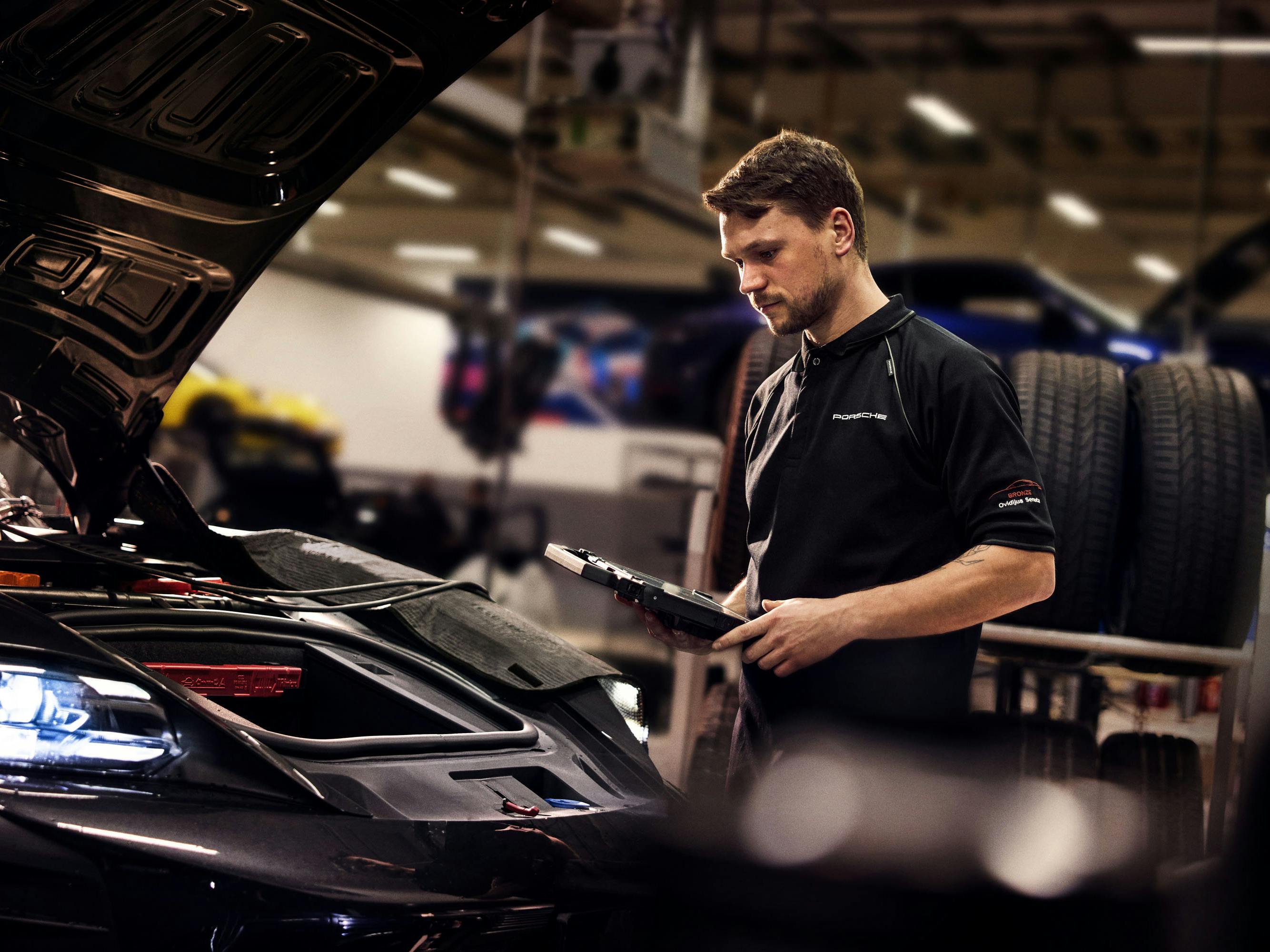 Porsche technician with diagnostic tablet stands beside open hood of Porsche vehicle in workshop.
