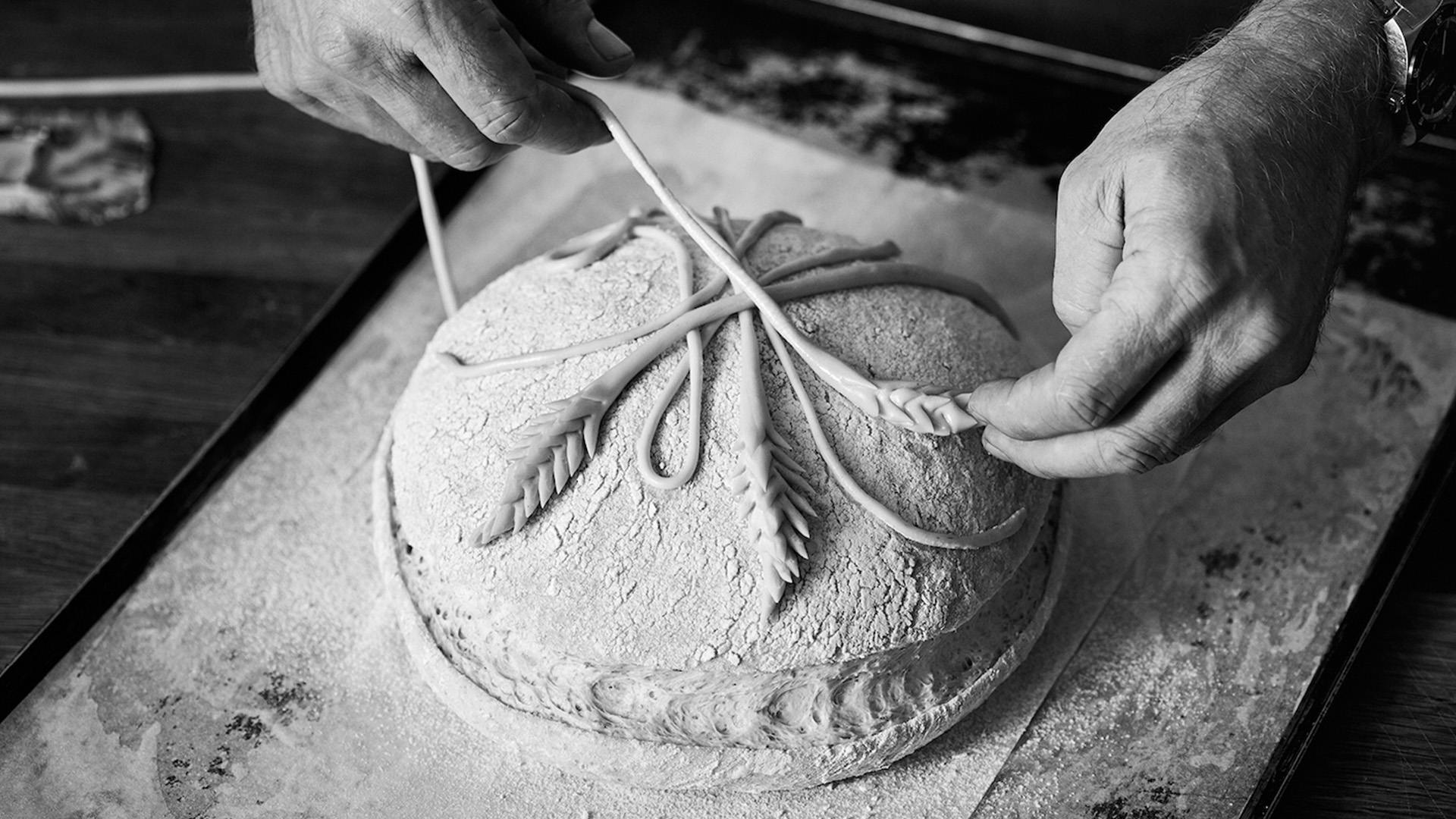 Baker decorates round bread dough with intricate dough strips and wheat patterns.