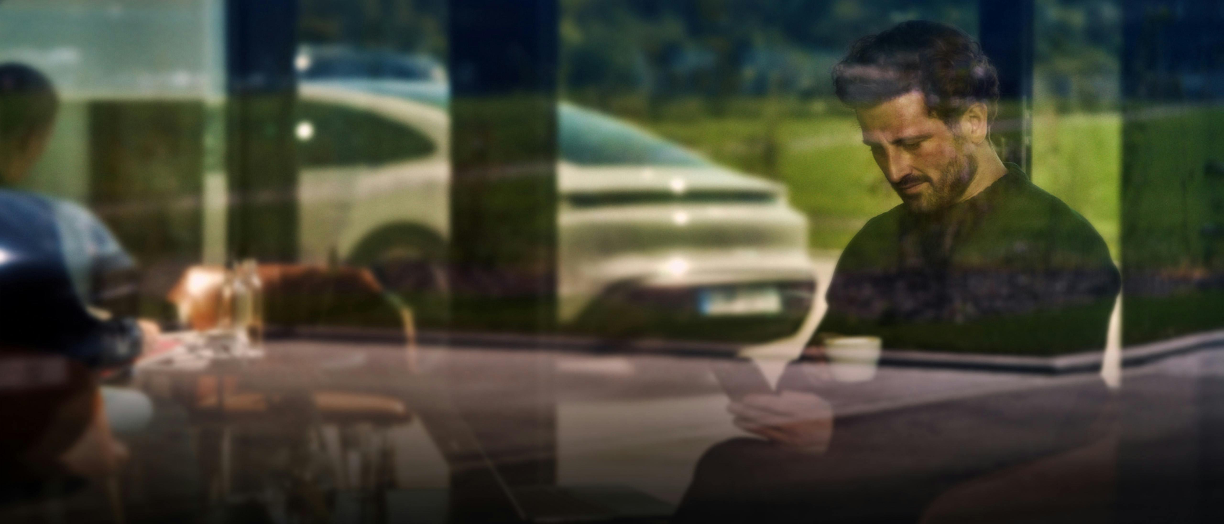 A Porsche car is visible through a glass window in the background, parked near a green outdoor area. Indoors, a person is seated at a table.