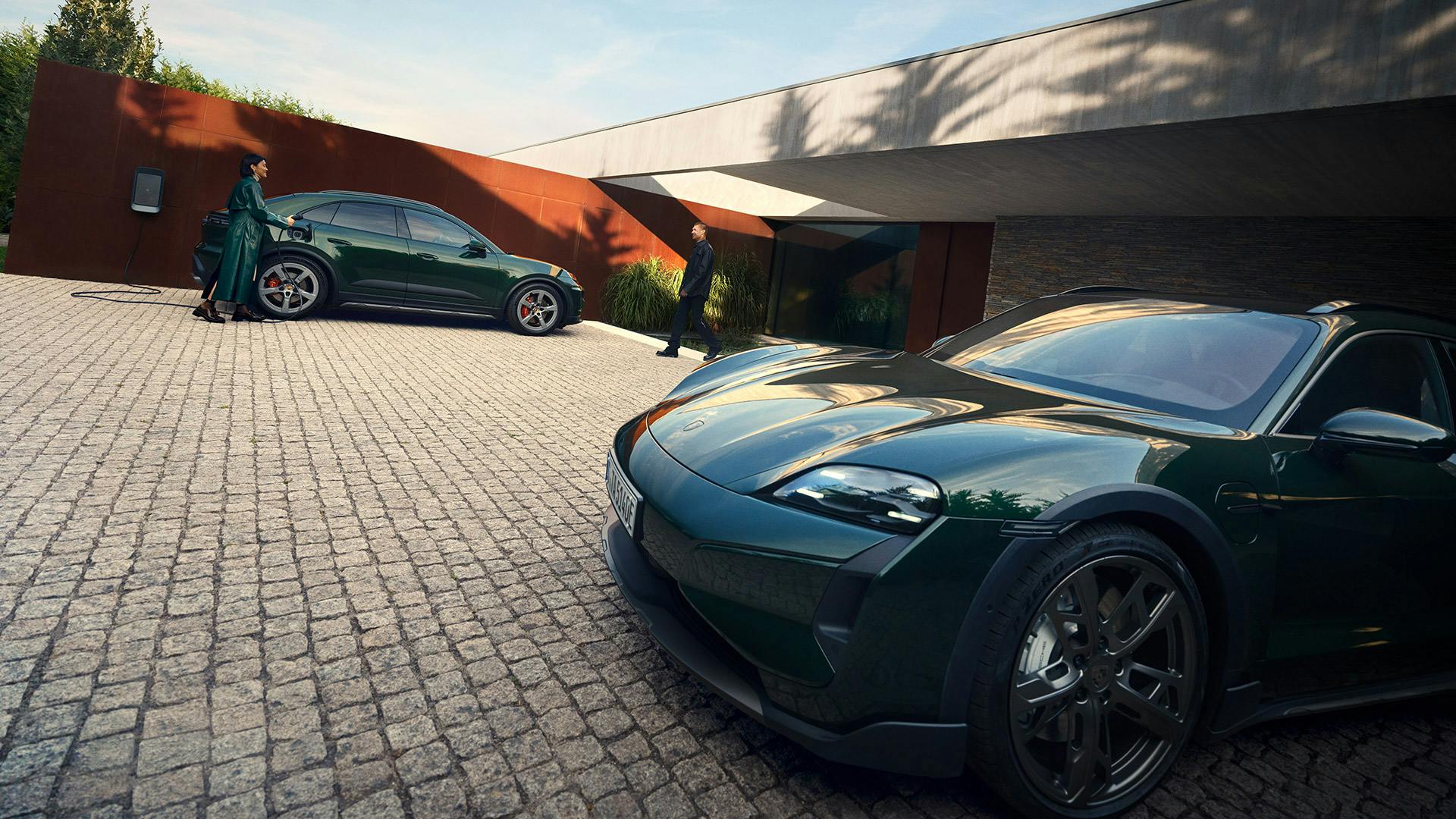 Deux véhicules électriques Porsche vert foncé sont stationnés dans l’allée d’une grande maison moderne.​