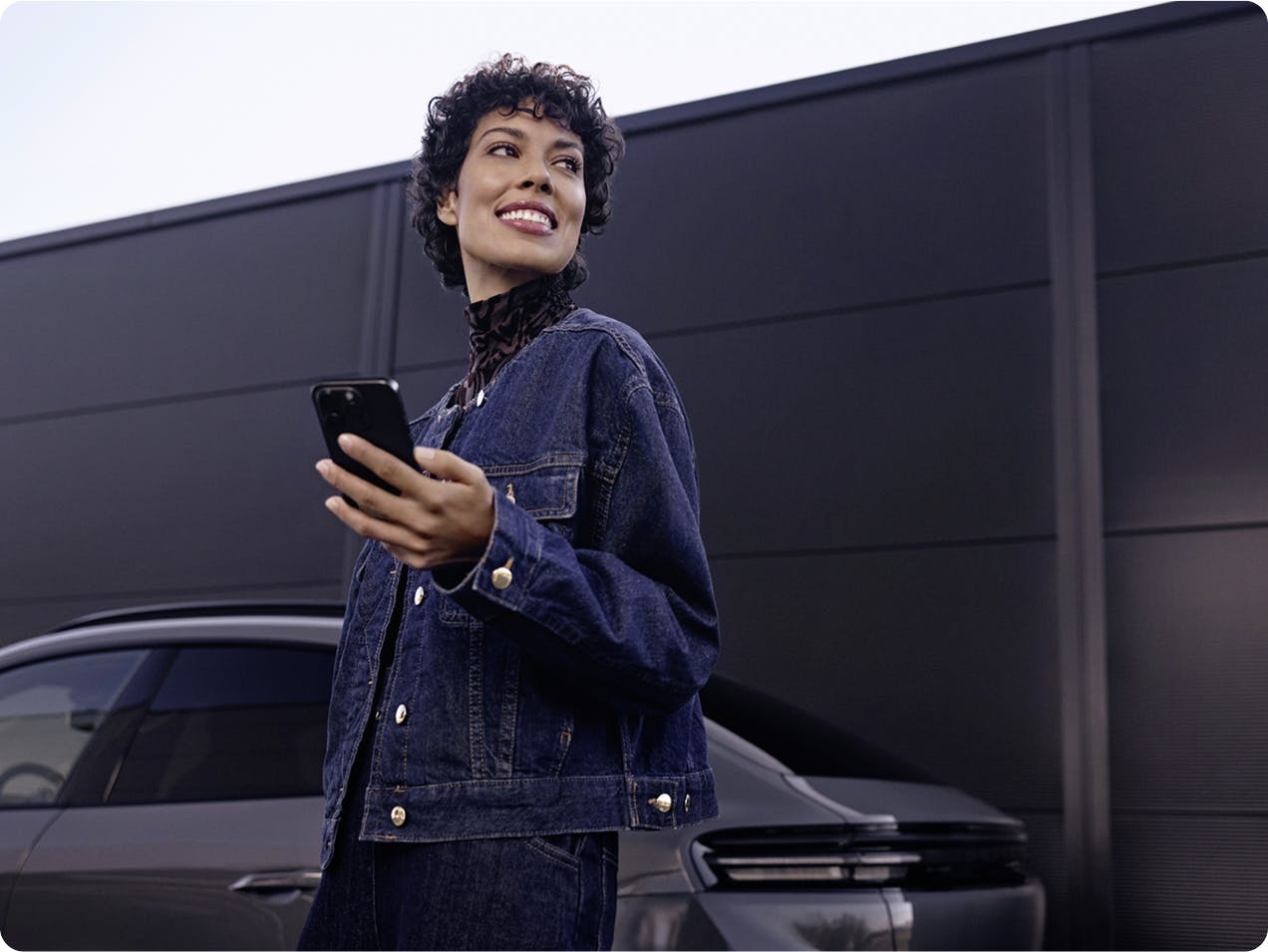A smiling woman with a smartphone in her hand stands in front of a Porsche Macan.