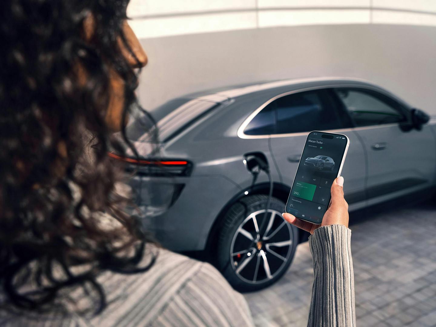 A woman is looking at her smartphone. A Porsche Macan can be seen in the background. A charging cable is plugged into it.