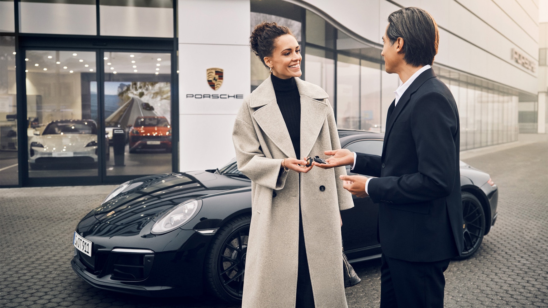 A woman receiving a car key from a man in front of a Porsche car and a Porsche center.