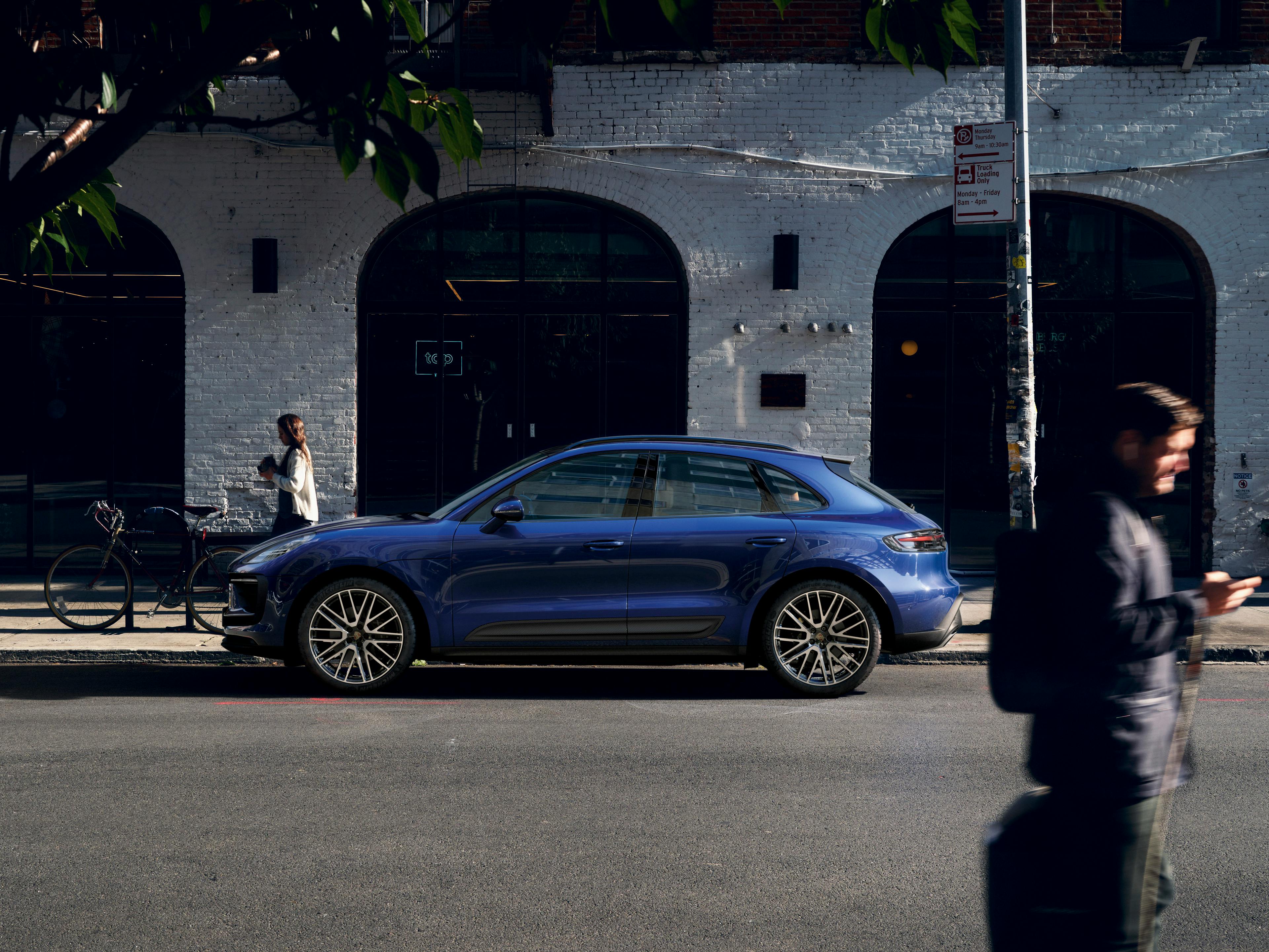 The Macan parked on the road, in front of a white building that looks like a shop, there is a man walking past in the foreground while a woman walking in the opposite direction in the foreground, nearer to the car.