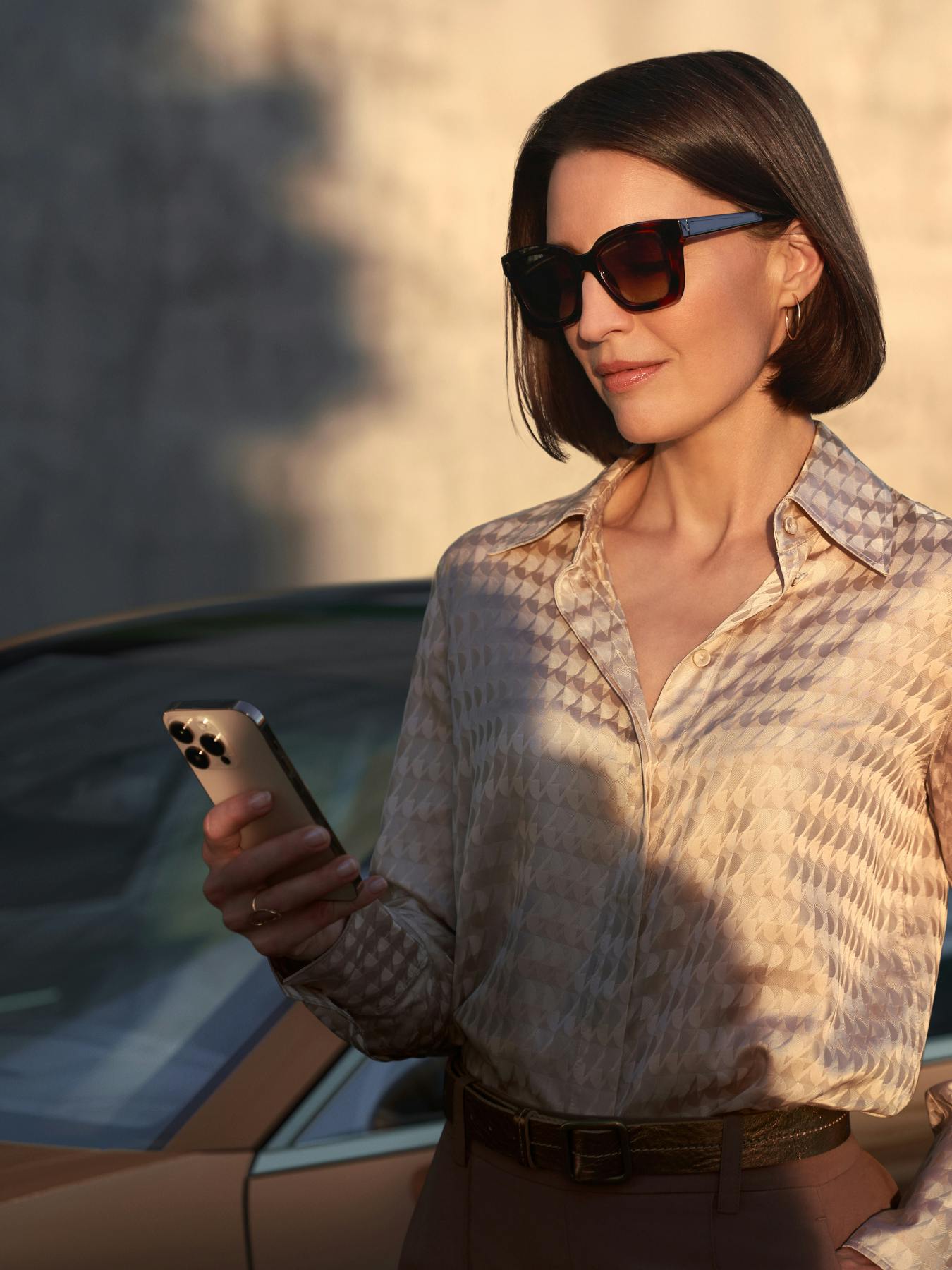 A woman with a dark brown bob stands in front of a Panamera Turbo E-Hybrid, looking down at a phone in her hand. She wears dark sunglasses, a patterned tan silk blouse, and a brown belt. The scene is lit by warm, low-angled sunlight, casting shadows on the wall behind her.