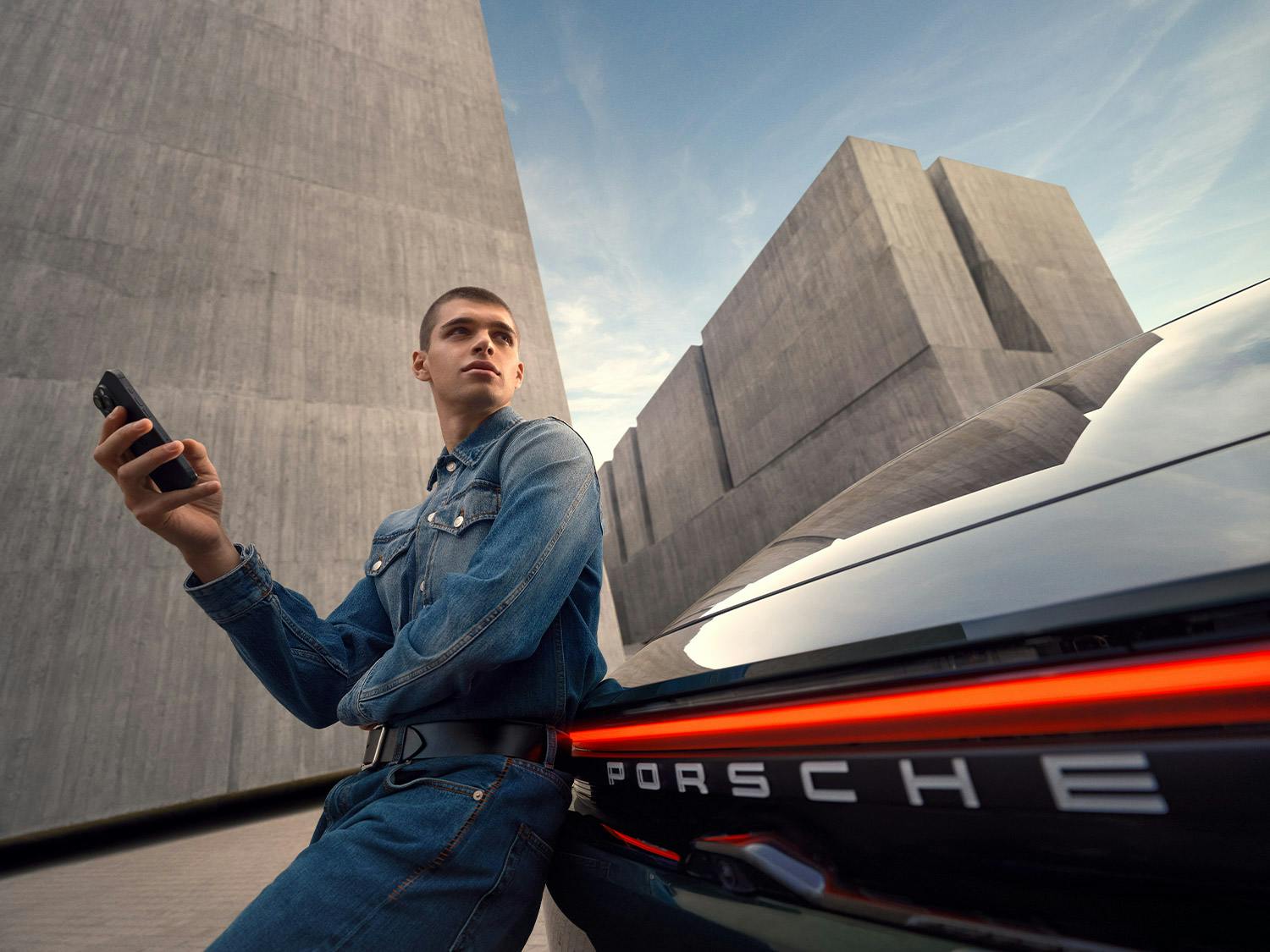 A person looking at their smartphone. A Porsche Macan 4S is charging at a Wallbox in the background.