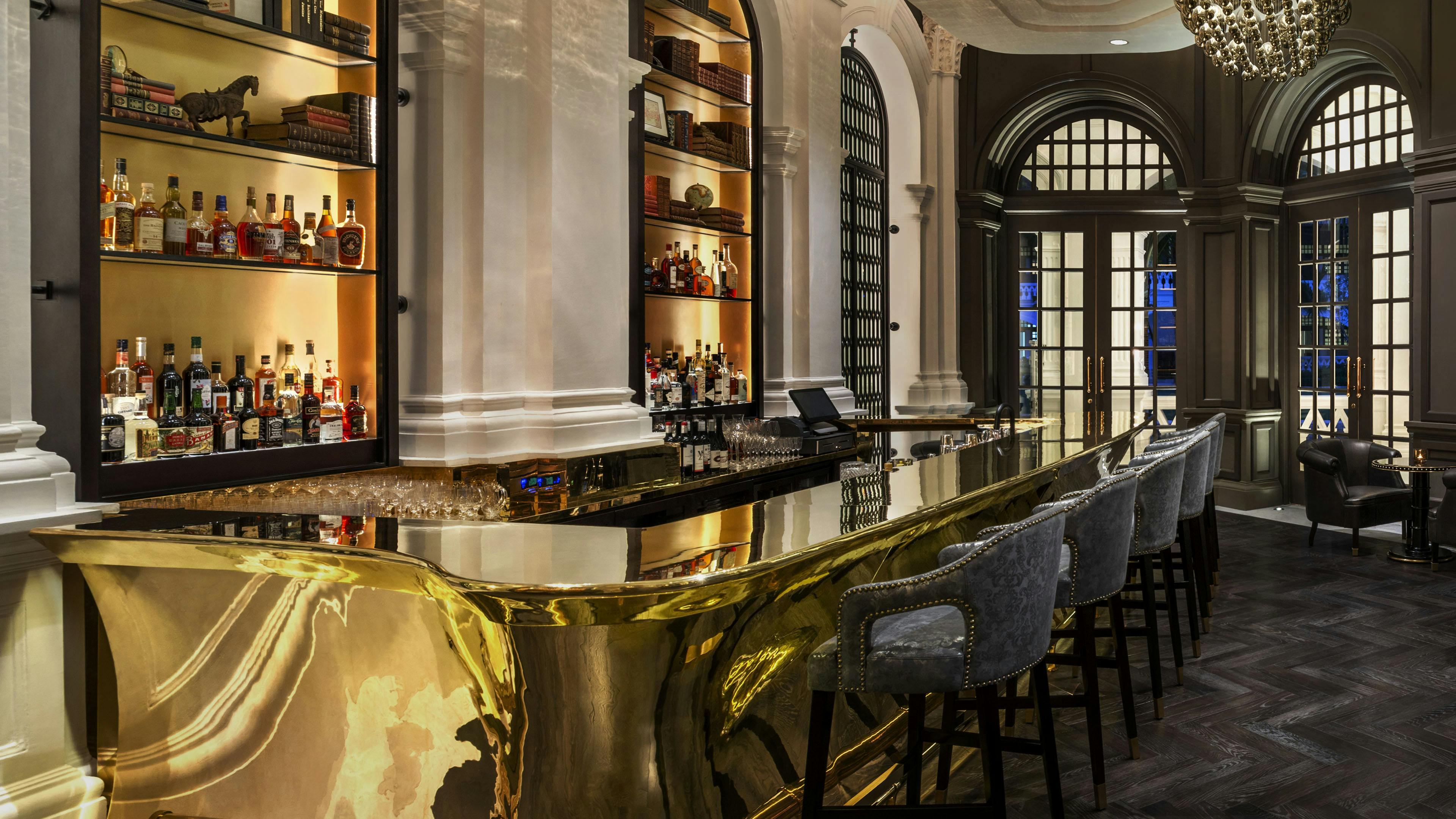 Elegant writers bar with gold counter, high-backed chairs, shelves of liquor bottles and books at Raffles Hotel.