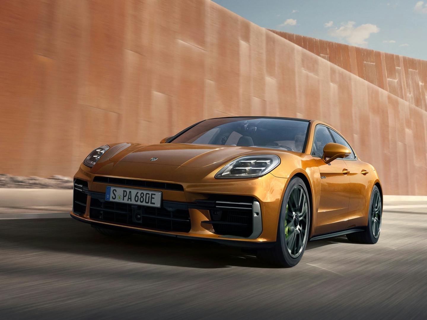 An orange Porsche Panamera Turbo E-Hybrid driving on a light-coloured, textured surface against a neutral, two-tone gradient background. The car is angled towards the left.