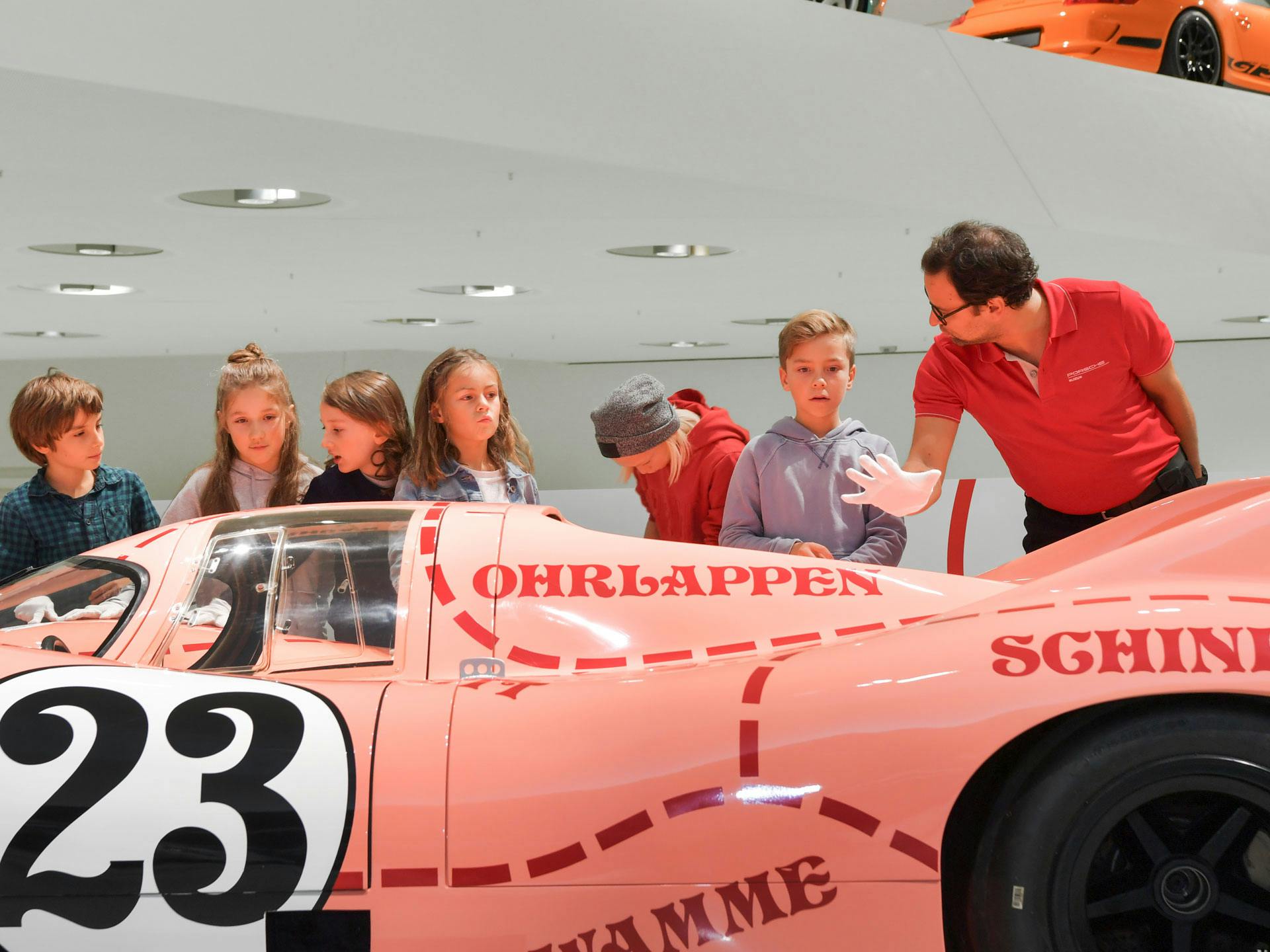 Eine Gruppe von Kindern vor einem rosanen Porsche-Exponat mit einem Museumsführer, der über das Auto spricht
