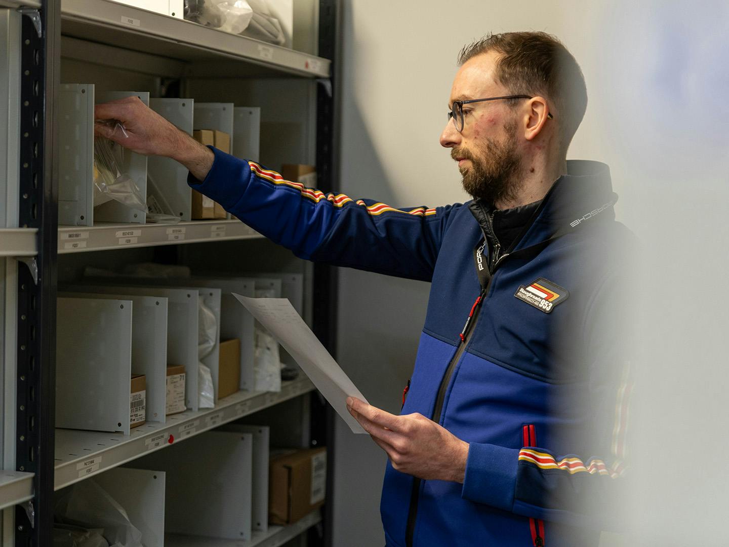 Un employé de Porsche se tient à côté d'une étagère et prend quelque chose dans un compartiment de l'étagère tout en tenant un morceau de papier dans son autre main.