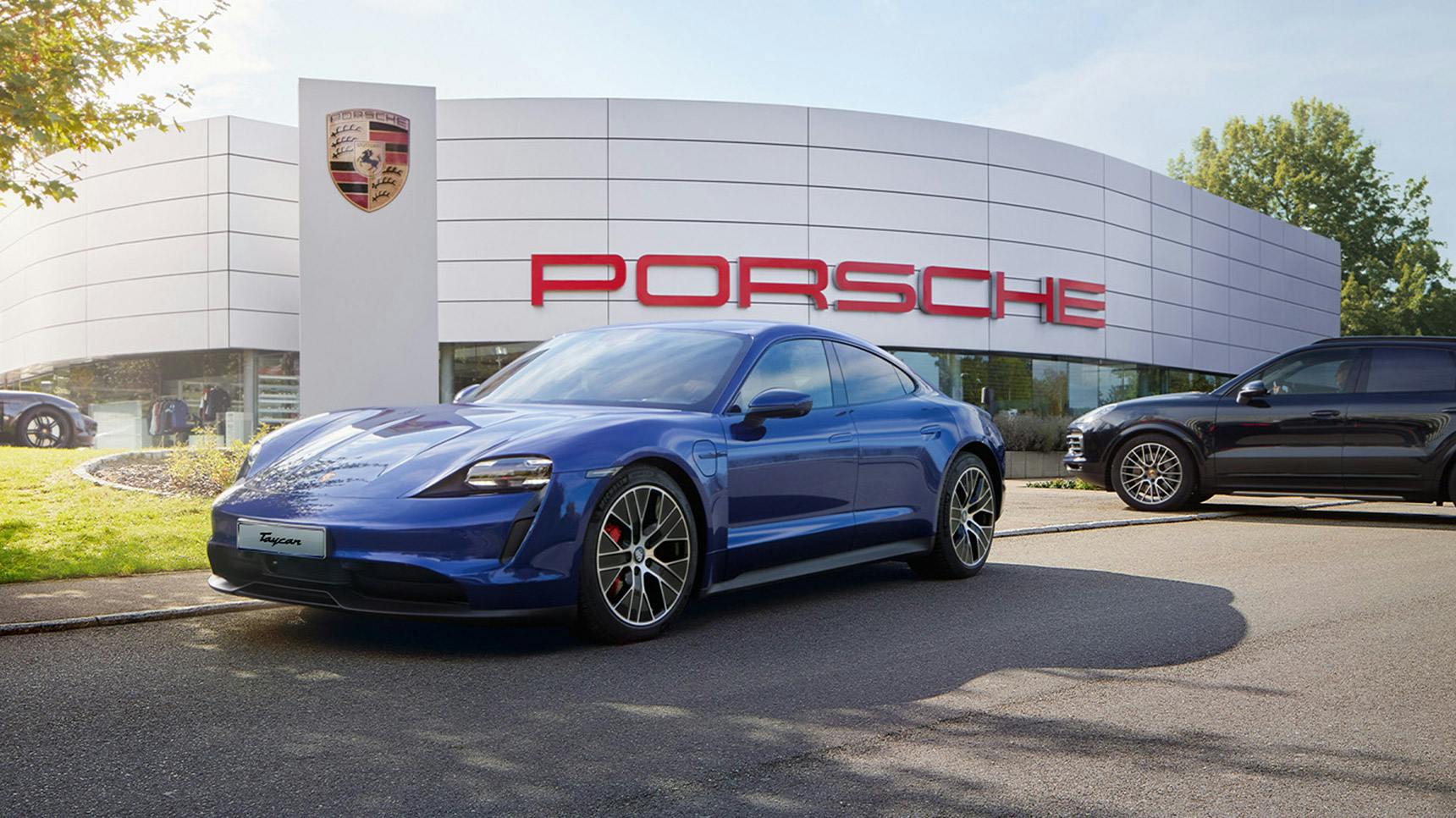 A blue Porsche Taycan is parked outside a dealership with the Porsche logo prominently displayed on the building.