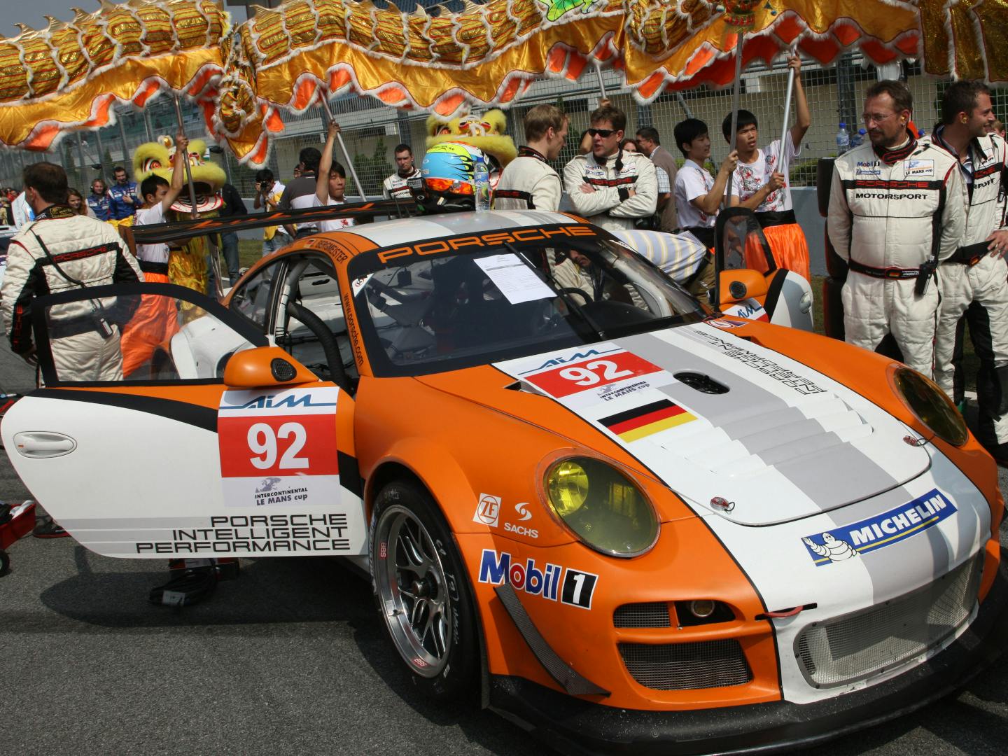 The orange and white Porsche 911 GT3 R Hybrid race car on the starting grid, surrounded by crew and onlookers.