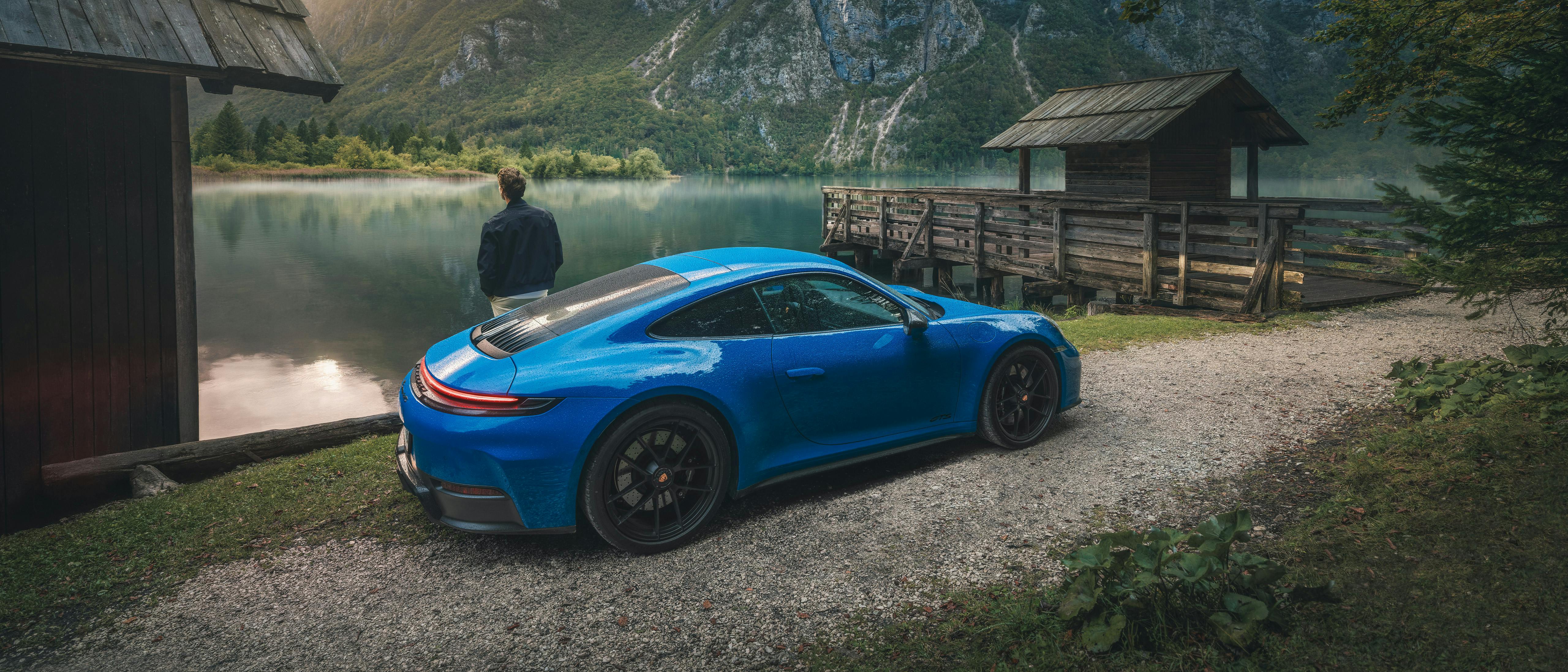 A man leaning on a blue Porsche 911 parked against a lakeside cottage with mountains in the background.
