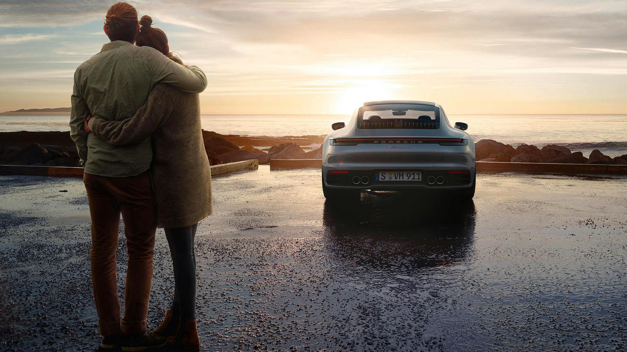 Una coppia si abbraccia affettuosamente al tramonto davanti a una Porsche 911, con il mare calmo sullo sfondo.