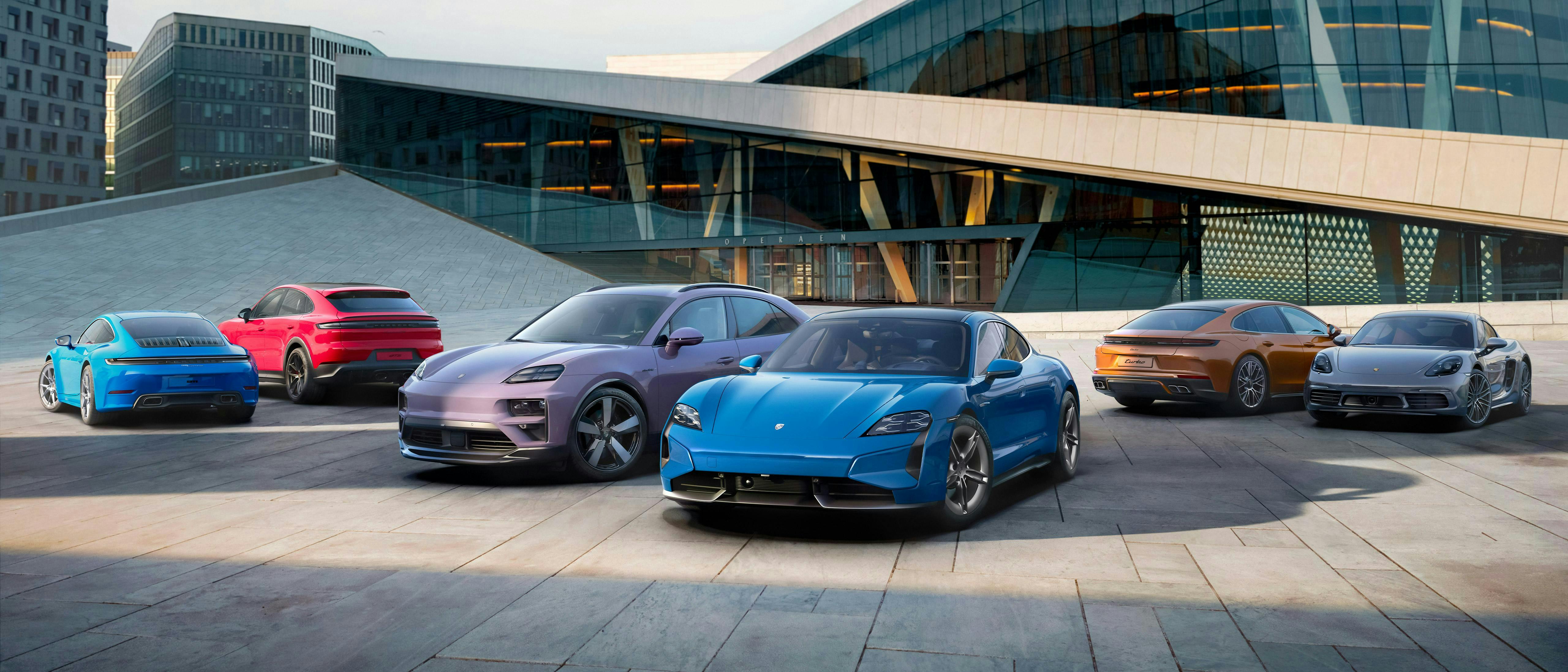 A lineup of Porsche cars, including SUVs and sports models, displayed in front of a modern architectural building under daylight.