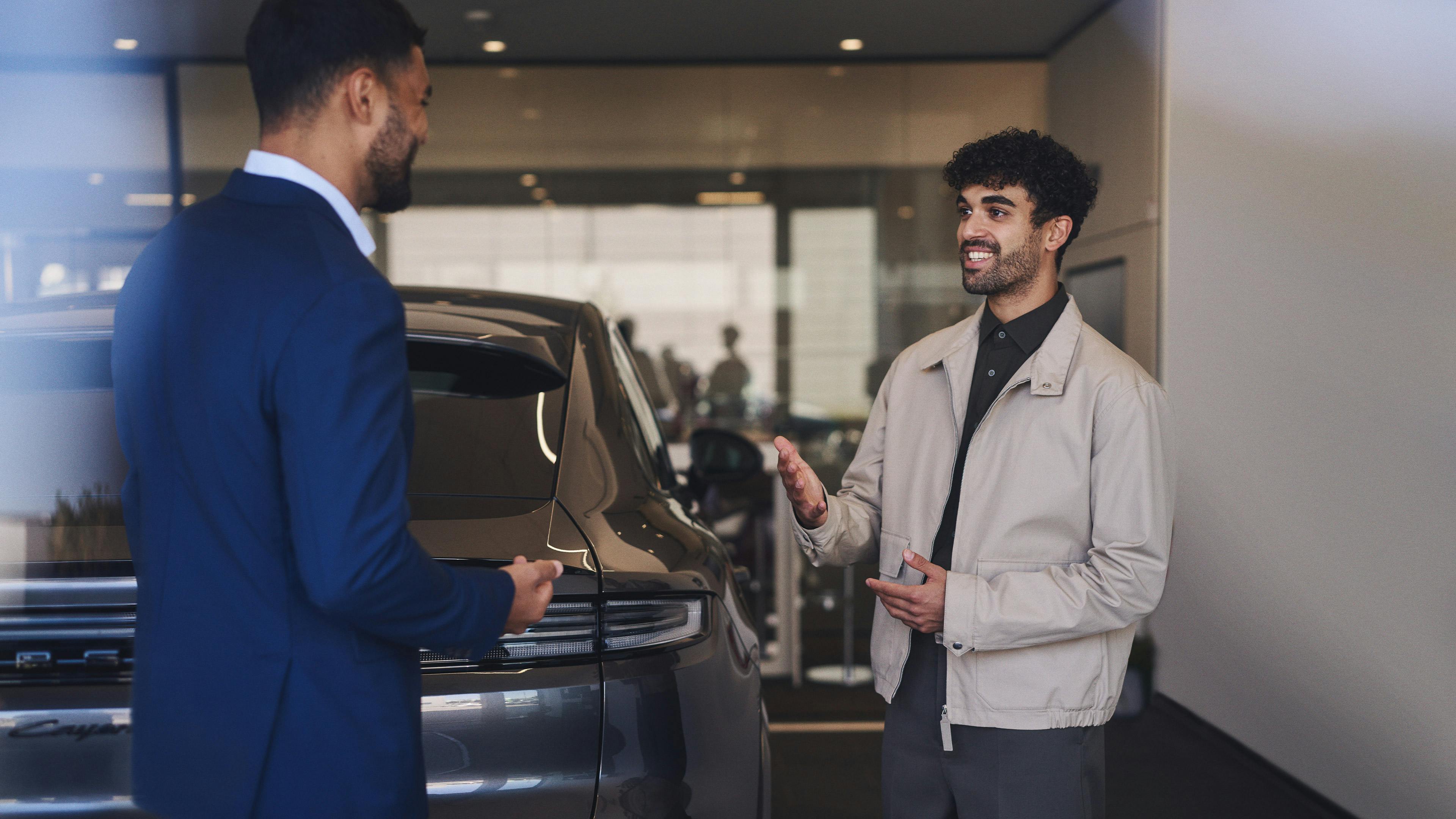 A Porsche employee in a blue suit and a customer talk in a Porsche showroom next to a Porsche.