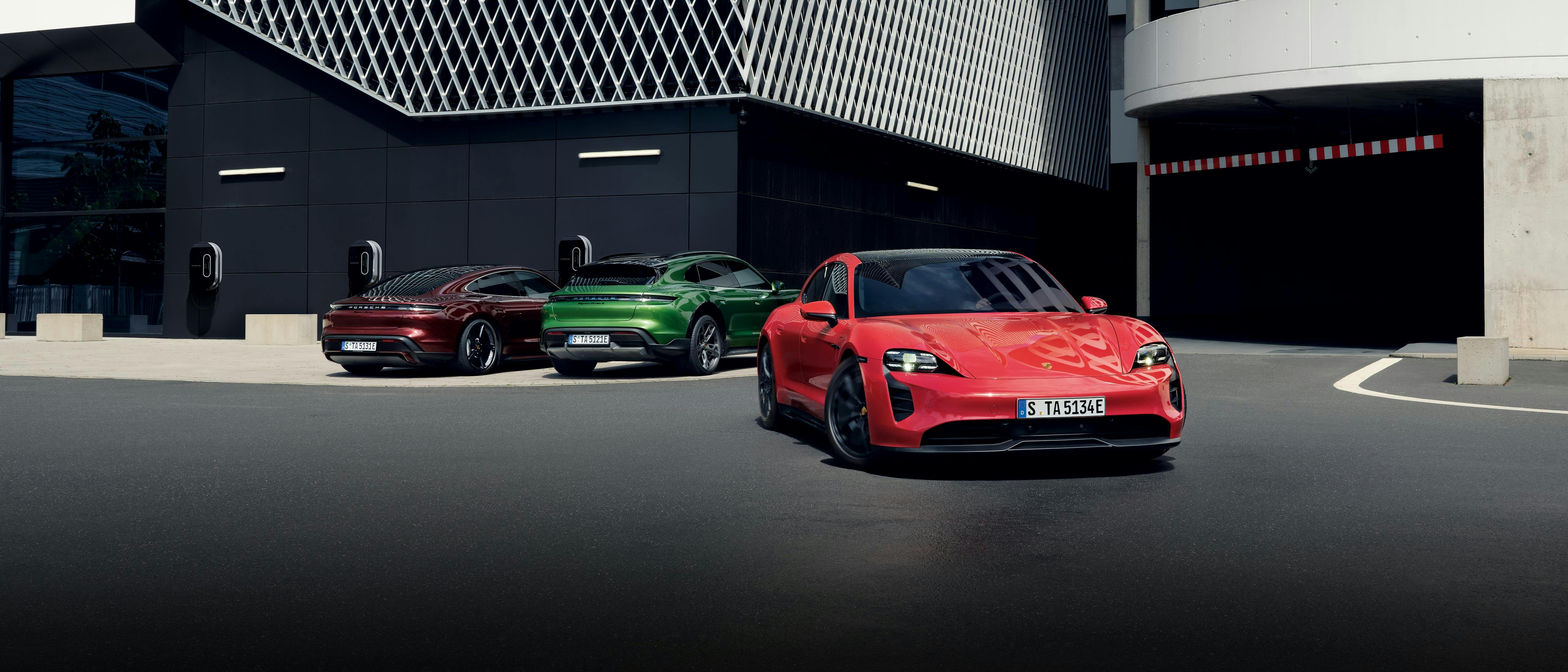 Trois modèles Porsche Taycan sont garés devant un bâtiment moderne. Des bornes de recharge sont accrochées au mur du bâtiment.