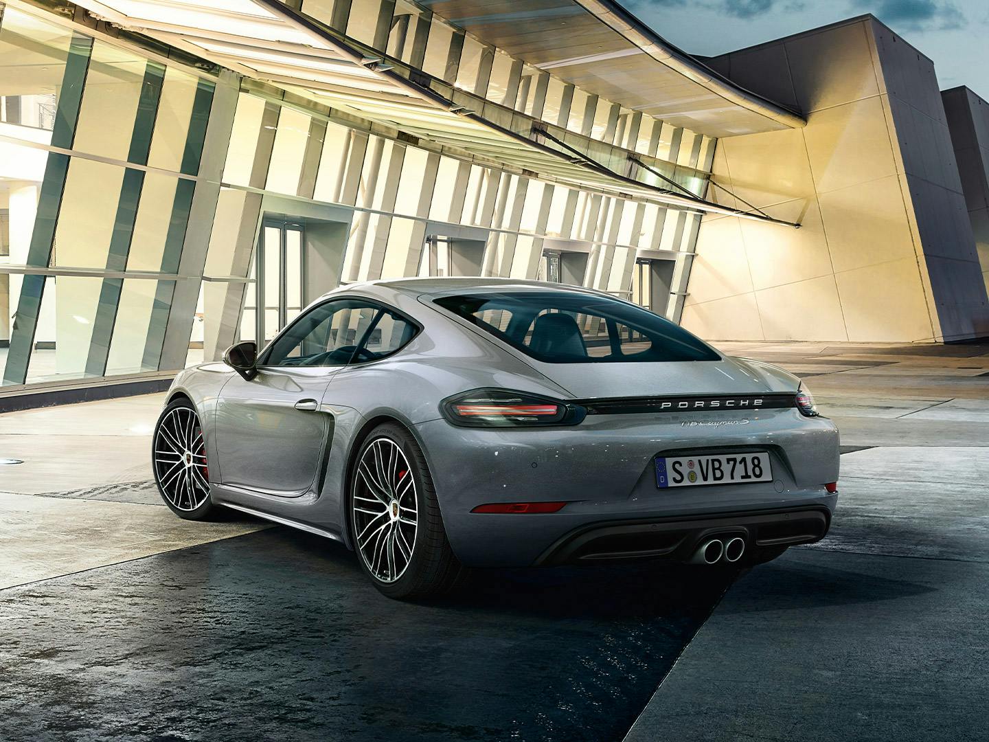A silver Porsche 718 Cayman in front of an illuminated building.