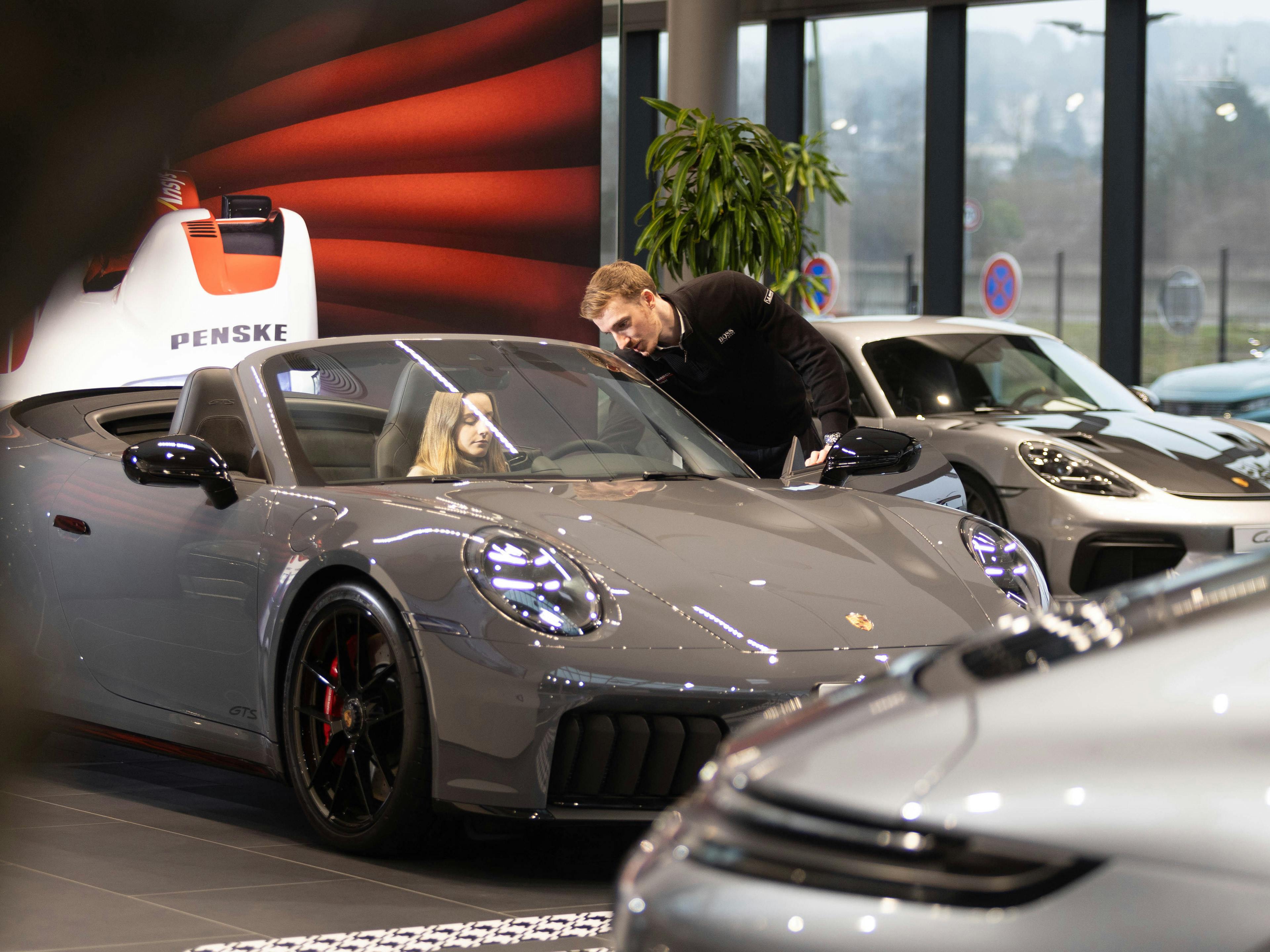 Un collaborateur de Porsche, debout à côté d'une Porsche grise au toit ouvert, parle à une cliente assise sur le siège conducteur du véhicule.