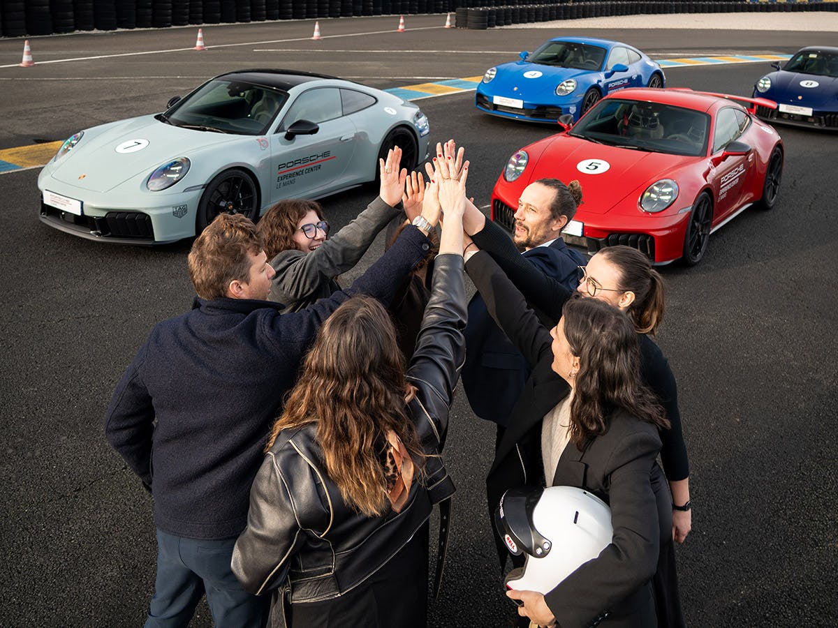 Sept personnes se donnant un high-five sur le circuit du Mans devant le Porsche  Experience Center, avec des voitures Porsche en arrière-plan.