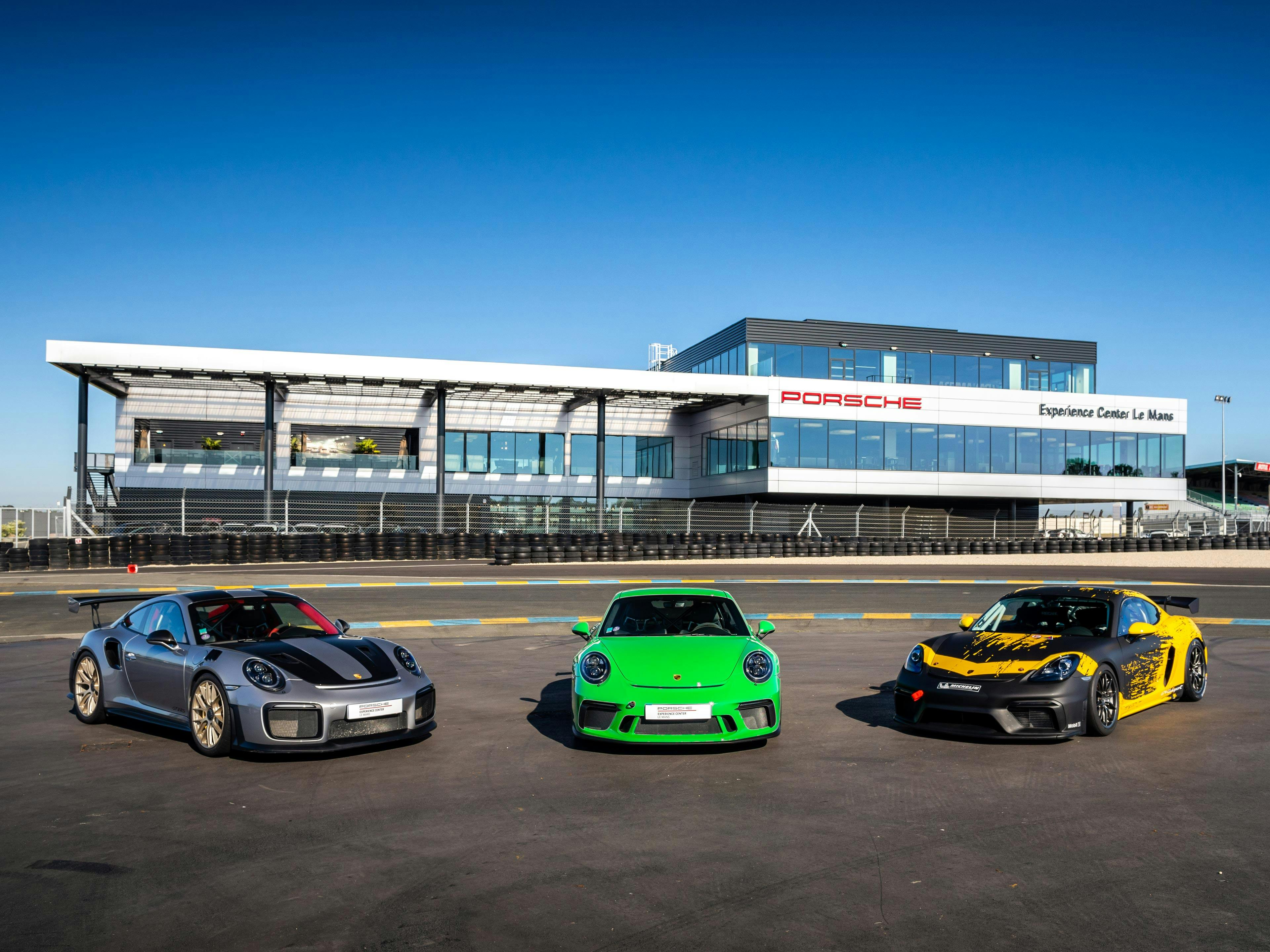 Tres vehículos Porsche estacionados en una superficie dentro del circuito frente al Porsche Experience Center Le Mans.