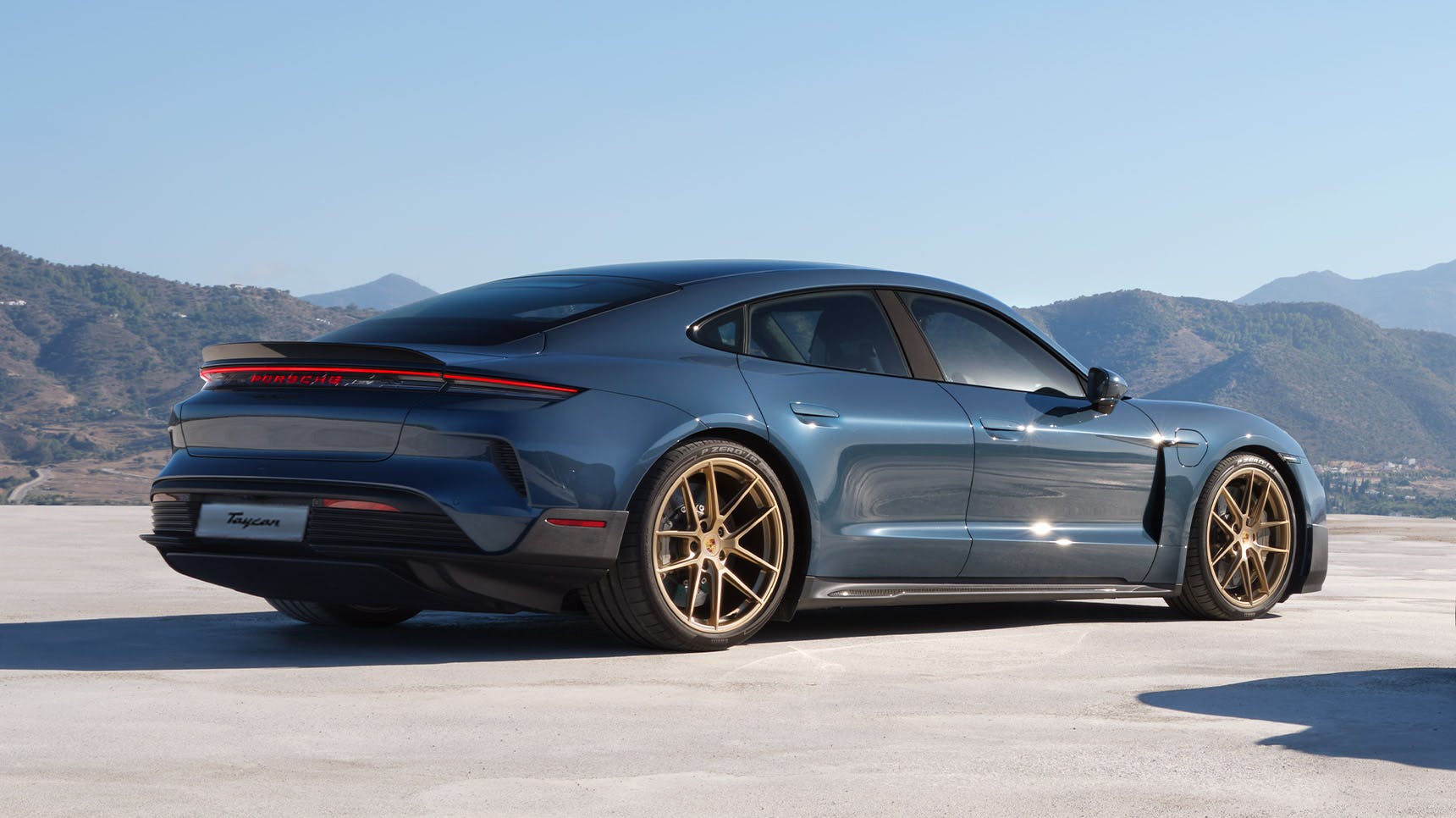 A blue Porsche Taycan with gold rims parked outdoors, showcasing its sleek design and sporty rear profile against a scenic mountain backdrop.