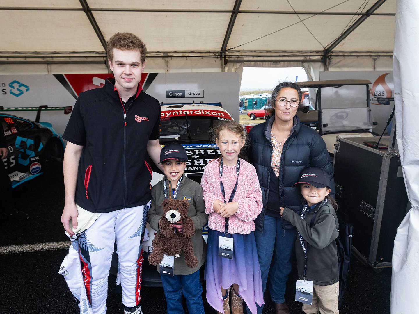 A group of five people – two adults and three children – standing outdoors in front of a Porsche race car at the Taupo Super400 event. Everyone thrilled and ready for an action-packed day.