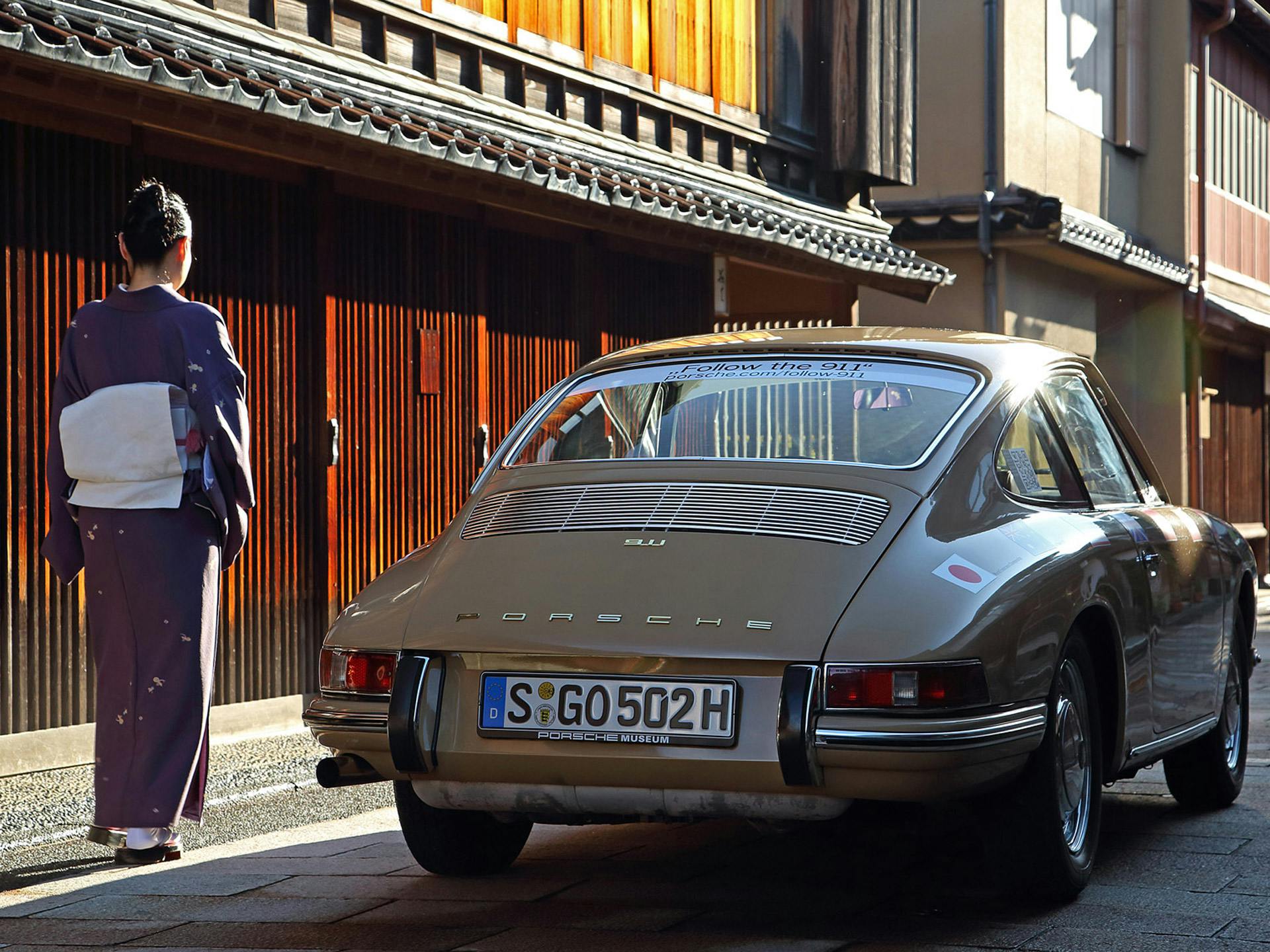 Heckansicht eines beigen klassischen Porsche auf einer Straße, neben Gebäuden und einer vorbeigehender Frau