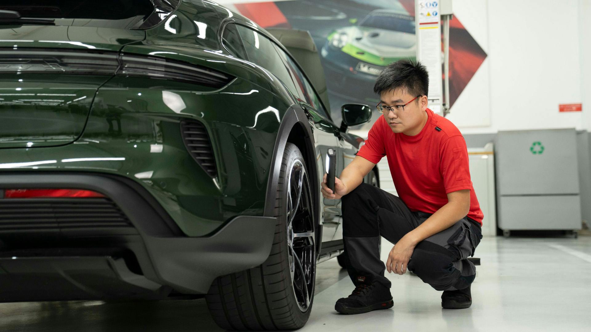 A technician in a red shirt inspecting the rear wheel of a dark green Porsche in a clean, brightly lit workshop.