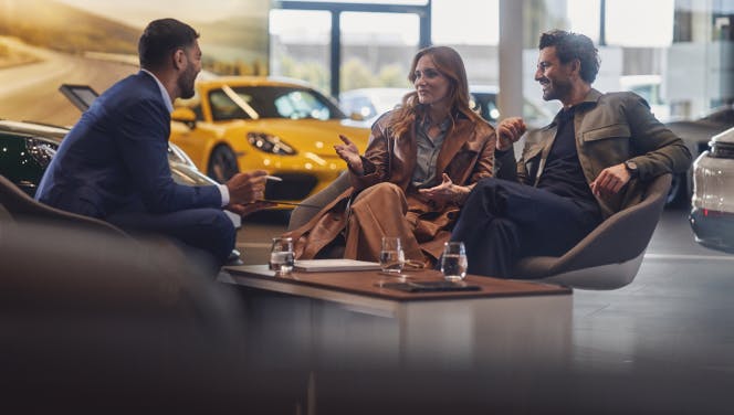 A couple is sitting opposite a Porsche employee in a consultation. There are various Porsche models in the background.
