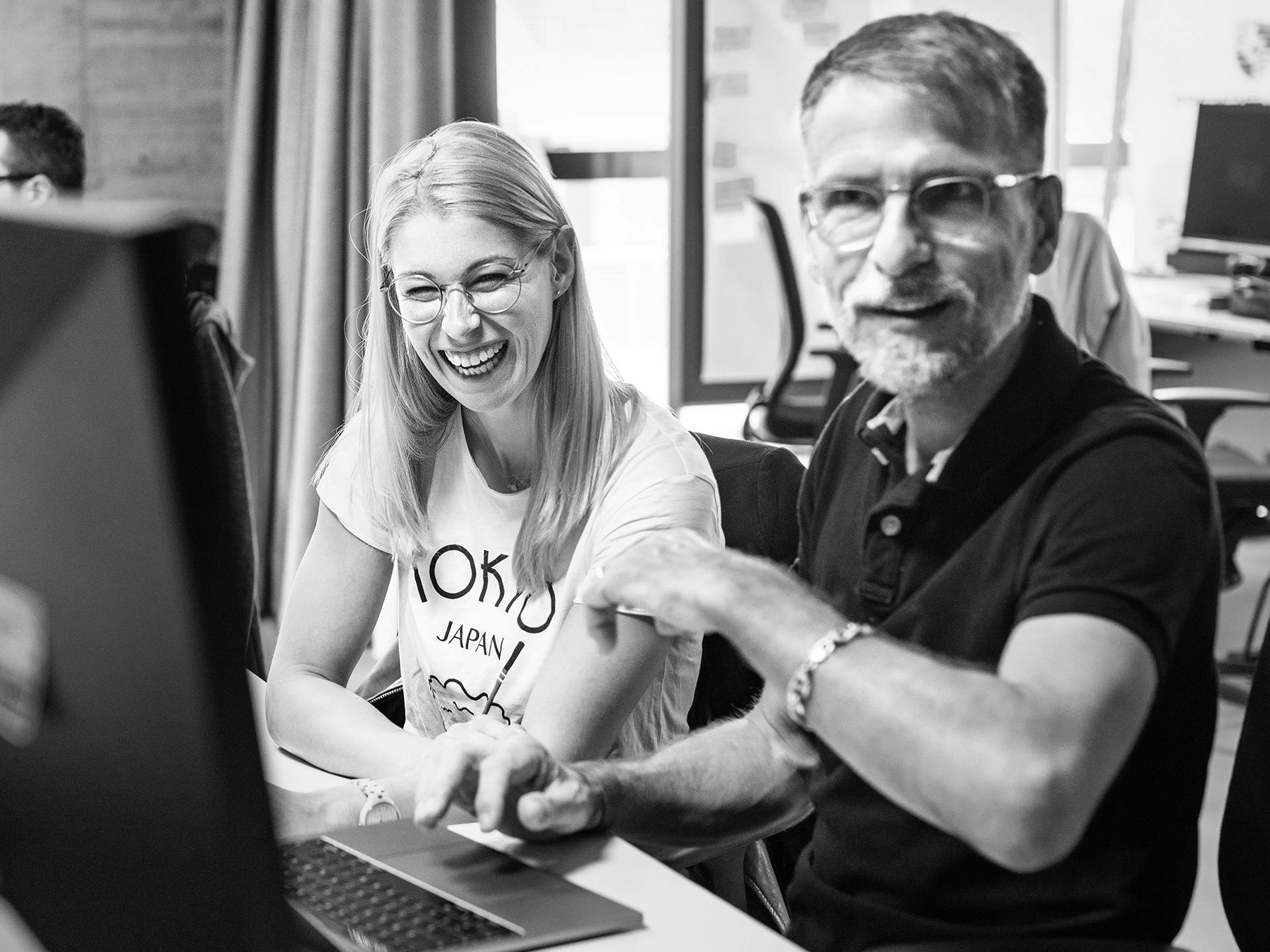 Black-and-white photo: two cheerful Porsche employees sit in front of a monitor.