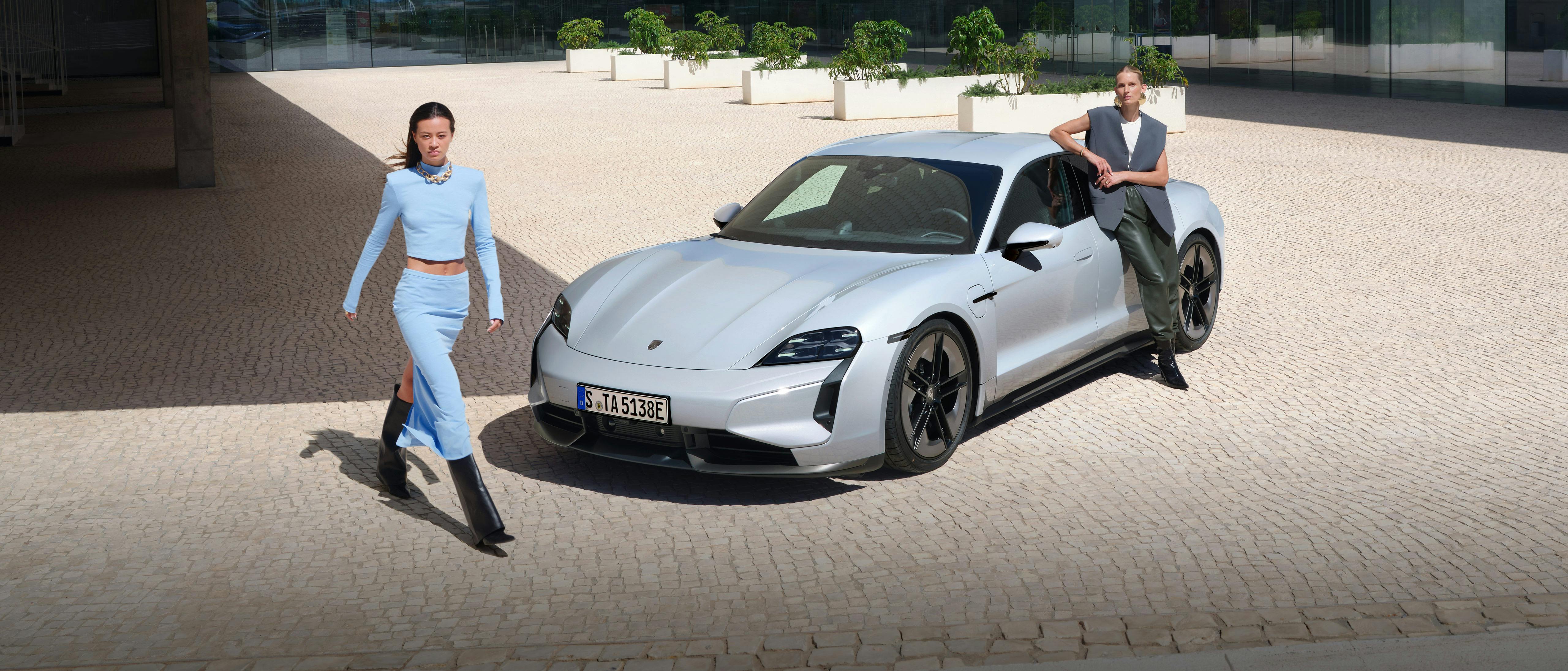 Ein grauer Porsche Taycan Turbo S steht auf einem weiss gehaltenem modernem Gebäude-Vorplatz. Zwei junge modern gekleidete Frauen schauen in die Kamera.