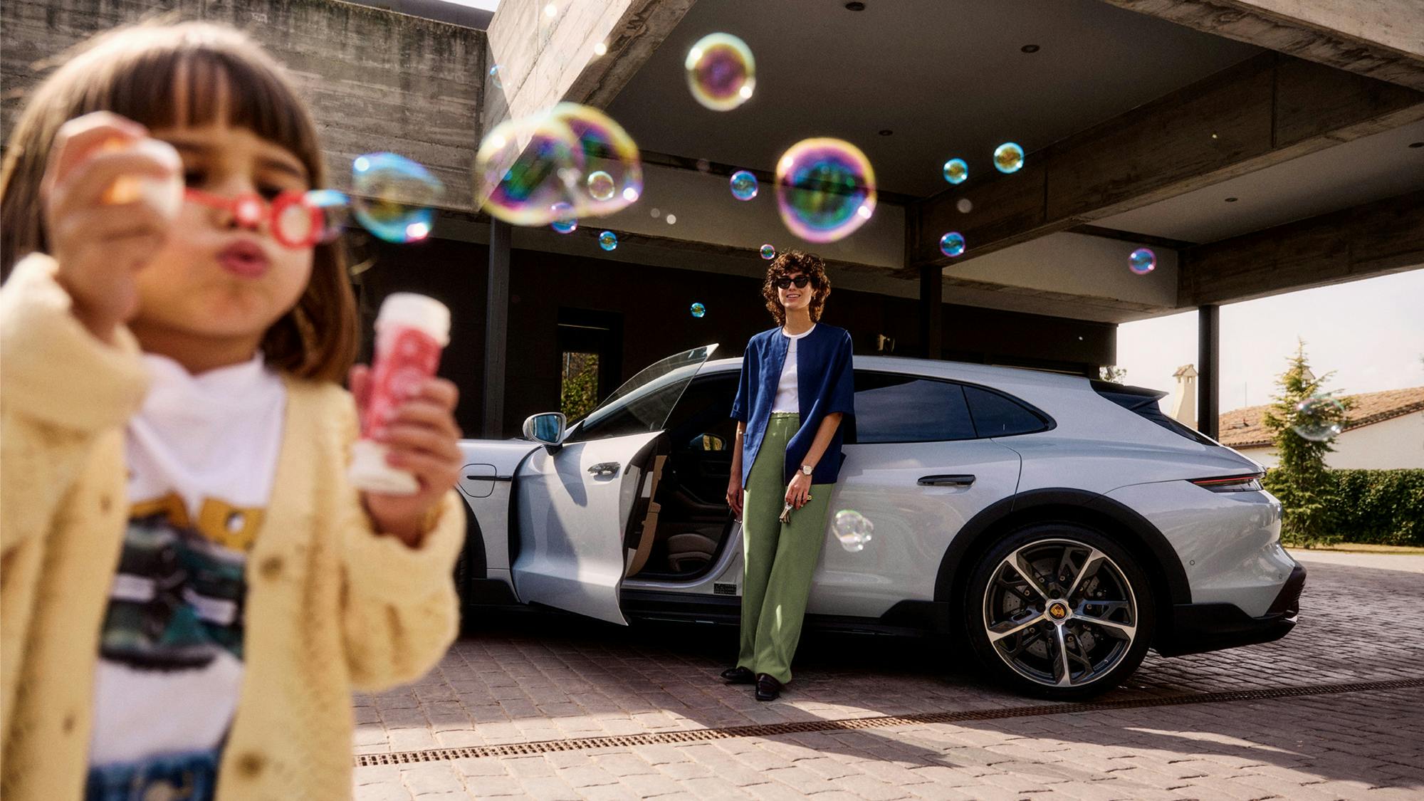 Frau mit Sonnenbrille lehnt lächelnd an einem silbernen Porsche Taycan Cross Turismo, während ein Kind im Vordergrund Seifenblasen bläst.