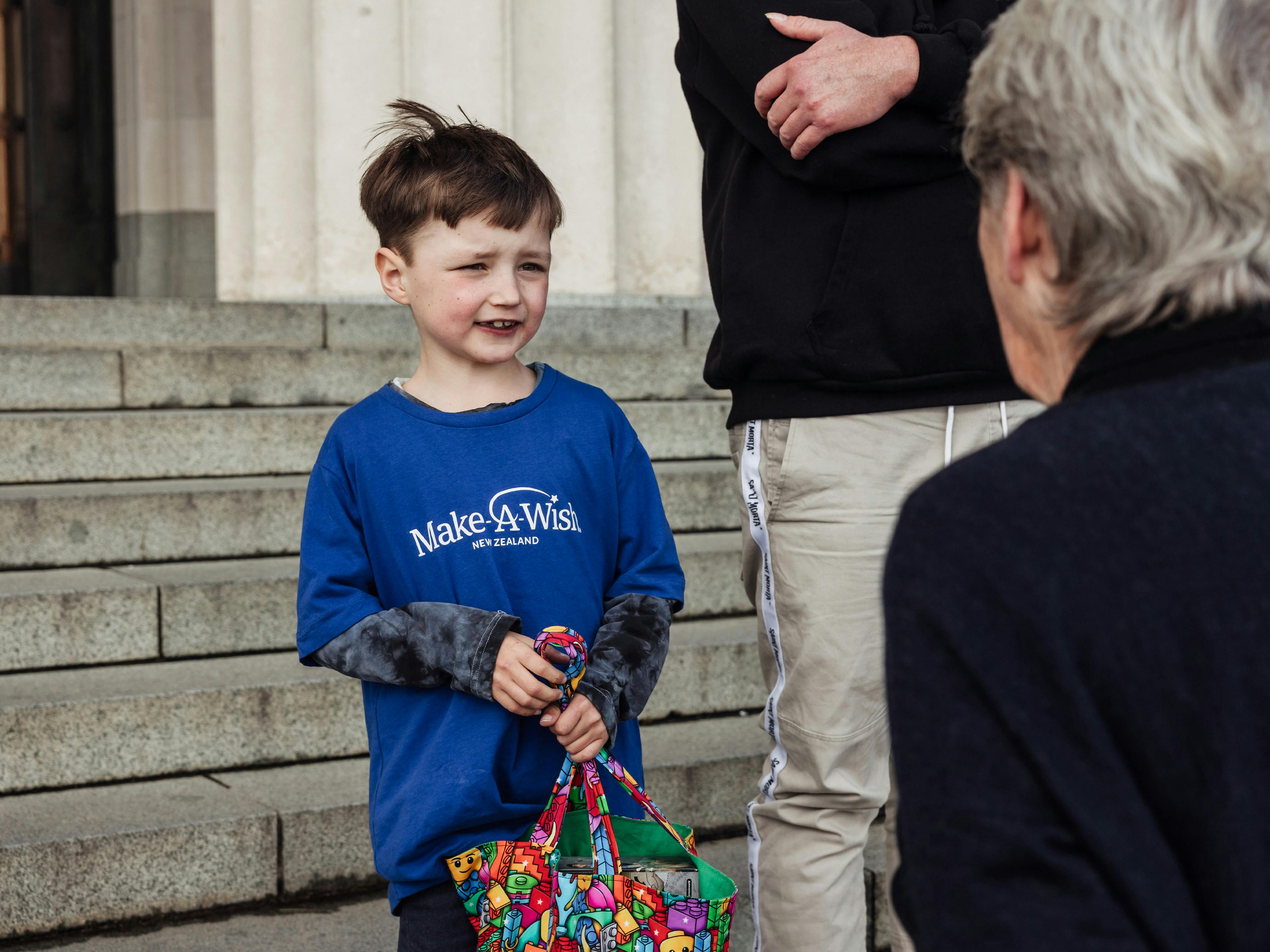 A young child wearing a Make-A-Wish blue long-sleeved shirt stands on stone steps, looking up towards an older adult whose back is to the camera.