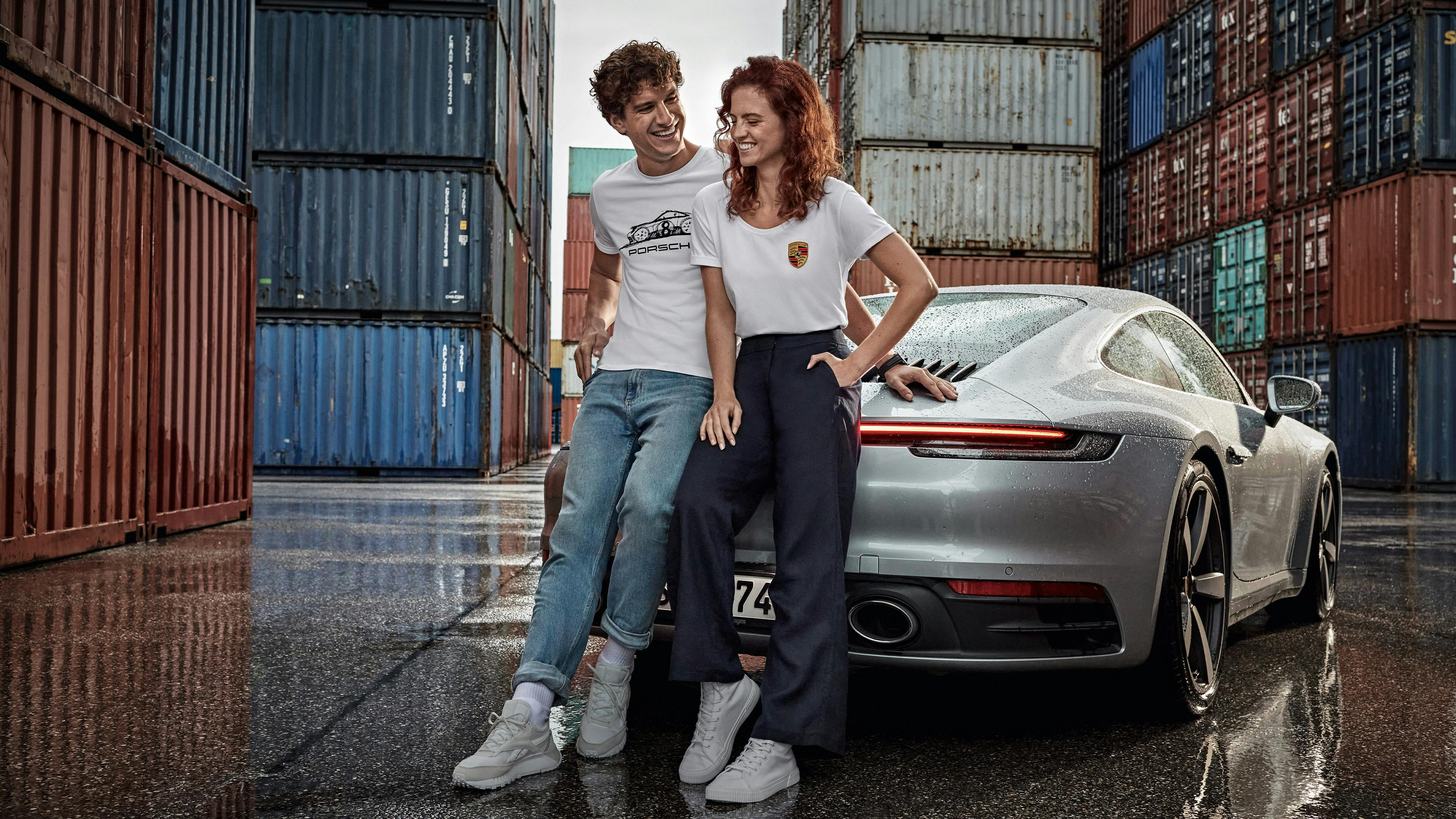 A smiling couple lean against the rear of a Porsche 911 in the middle of a container port.