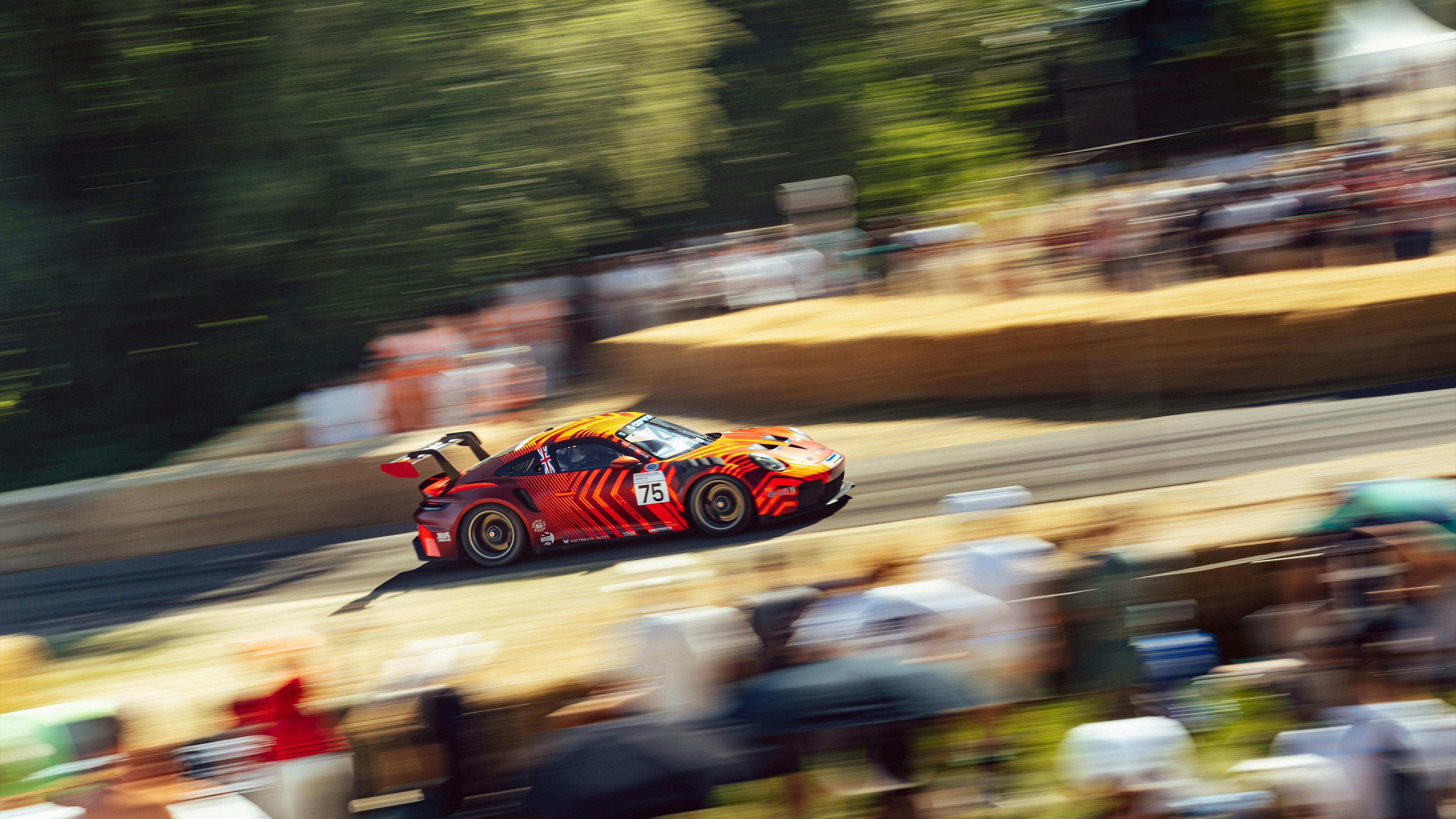 Orange and black liveried Porsche 911 GT3 racing up the Goodwood hillclimb, captured with motion blur against a backdrop of trees and spectators.
