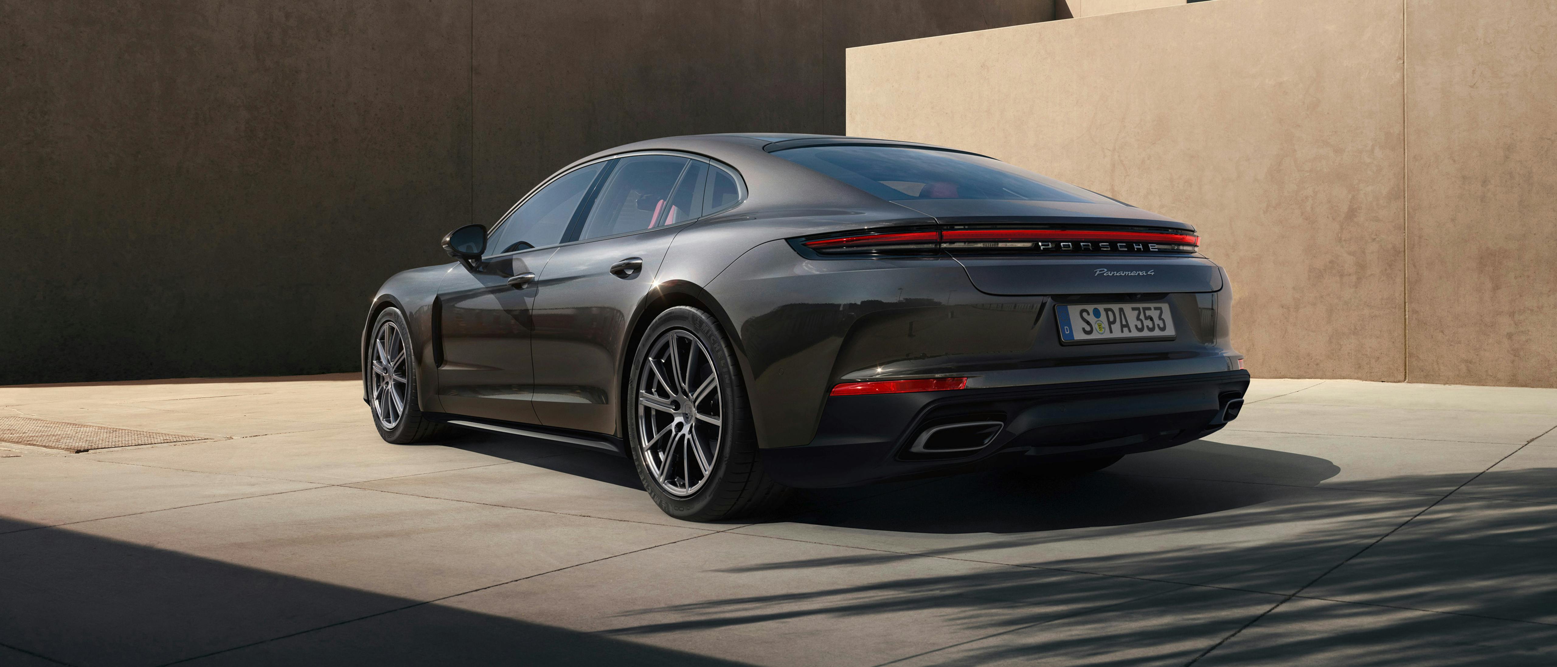 A dark grey metallic Porsche Panamera 4 parked in a modern, sunlit courtyard with minimalist tan-coloured concrete walls.