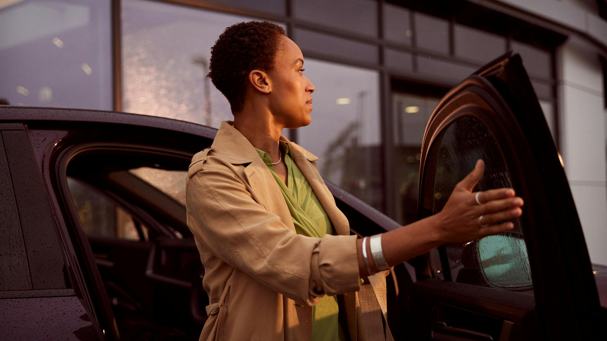 A person with short, dark hair stands by an open car door, looking toward the right.The background features a building with large glass windows reflecting the warm, golden light of late afternoon. Light raindrops are visible on the car's window and exterior.