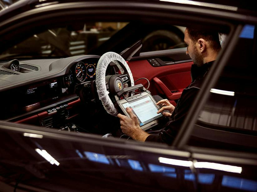 : A male technician sits in the driver's seat of a Porsche with a red and black interior. He is using a rugged diagnostic tablet connected to the car's systems. The steering wheel is covered in a protective plastic wrap, and the digital instrument cluster and center console screen are illuminated.