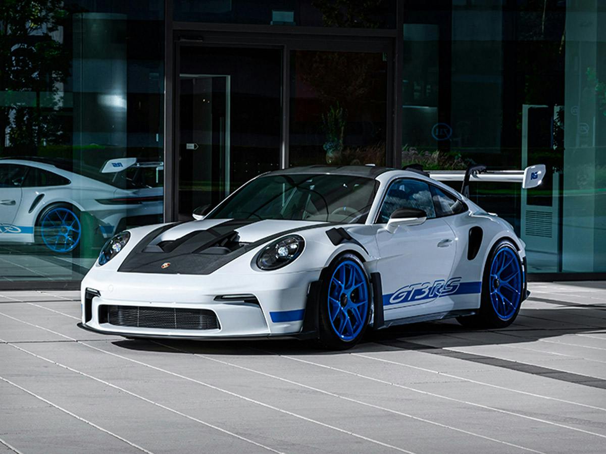 911 GT3 RS in Paint to Sample colour Purewhite with Sharkblue individualized wheels, shown from a front-side angle, parked in front of a modern glass building with a reflecting pool.
