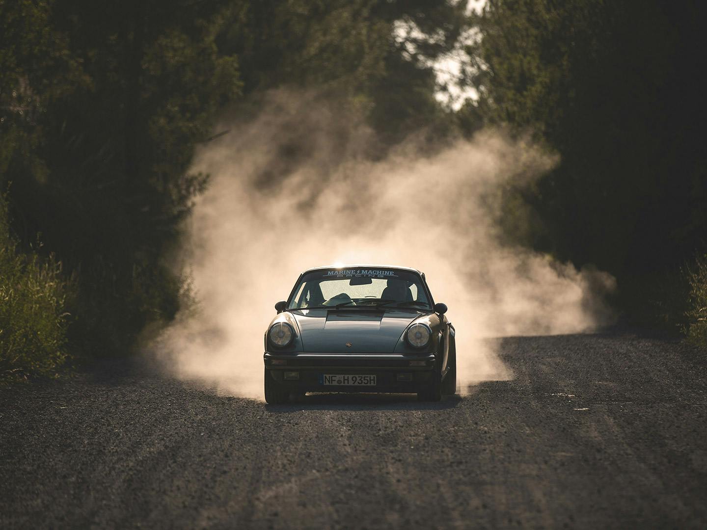 A classic Porsche sports car driving along a sunlit gravel road through tall trees, a dramatic plume of dust trailing behind as warm late afternoon light casts long shadows and highlights the car’s curved silhouette and chrome details; a partially obscured windshield banner is visible.